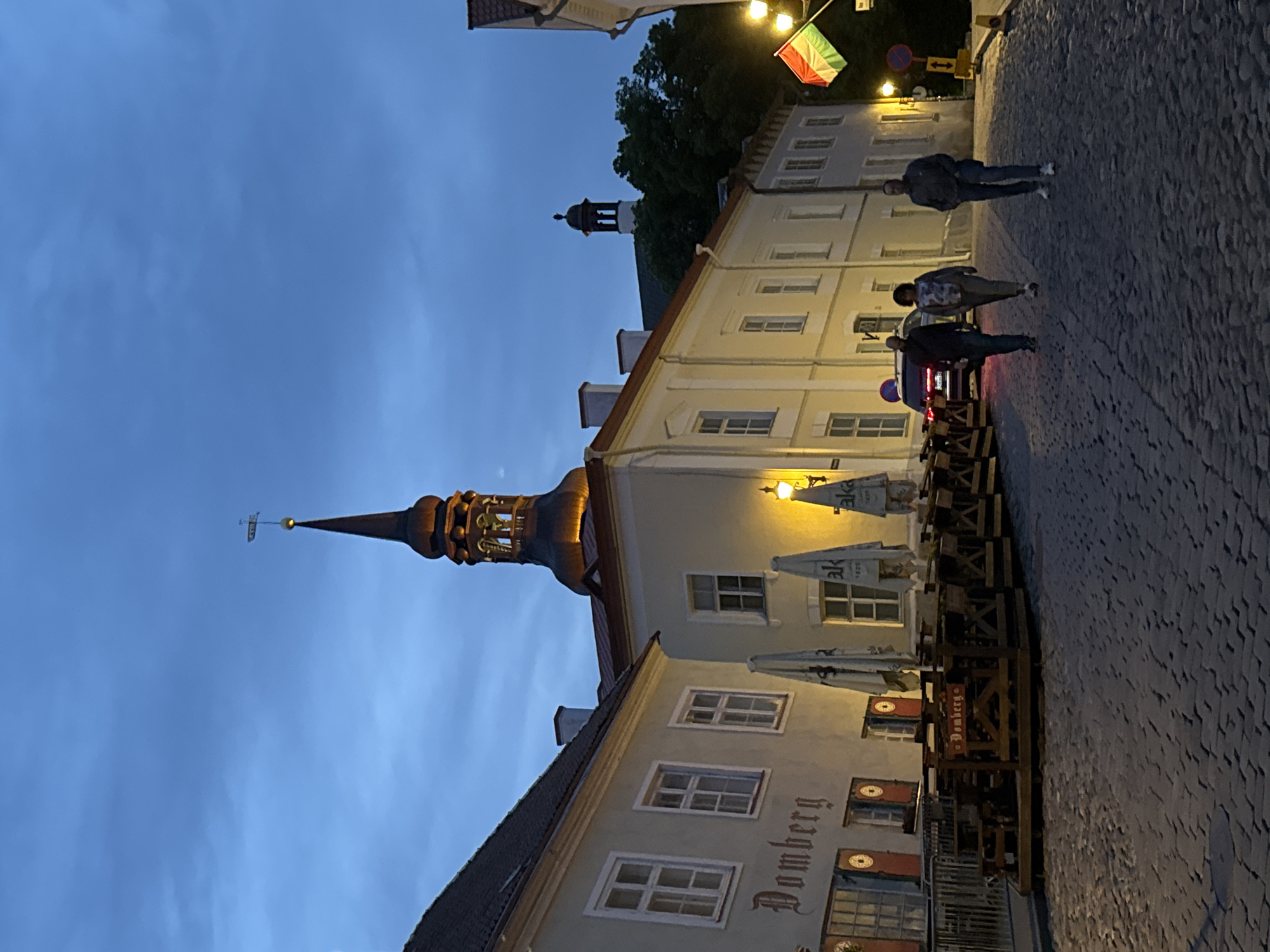This image captures a street scene in Tallinn, Estonia, at twilight. The architecture suggests a historic district, possibly near the Old Town. Prominent features include a church spire with a weather vane, illuminated by warm light, and a darker, more distant tower. Several buildings line the cobblestone street, displaying traditional facades with multiple windows. One building has a sign with "Domberg" in Gothic script. Outdoor seating areas with wooden tables and chairs, some with umbrellas, are set up in front of a restaurant or cafe. Three people are walking away from the camera down the street. A car is parked further up the road. A flag with horizontal stripes of green, white, and red is visible on a pole. Streetlights cast pools of light on the cobblestones. The sky is a deep blue, indicating the transition from dusk to night.