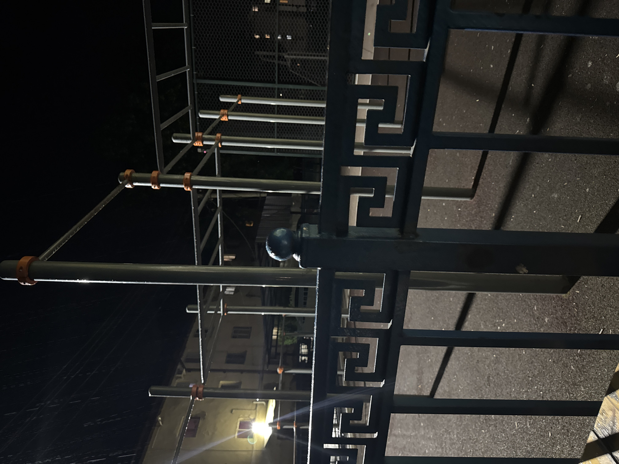 An outdoor exercise equipment area is depicted at night. A dark blue metal fence with a Greek key pattern is in the foreground. Behind the fence, there are several metal poles and bars of exercise equipment, some with orange accents. The ground is a dark asphalt surface. In the background, a building with illuminated windows is visible, and faint streaks of light suggest rain or moisture in the air. The overall scene is dimly lit by artificial light, creating a somber atmosphere.