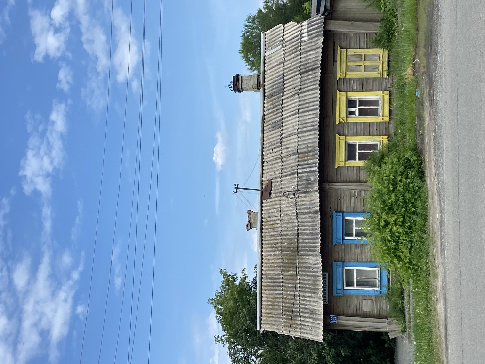 The image shows a weathered, wooden house with a corrugated roof in the city of Tyumen, Russia. The house has several windows, some with blue frames and others with yellow frames. A street sign with the inscription "Димитрова 102" is visible above two blue-framed windows. Dense green foliage surrounds the lower portion of the house. Power lines stretch across the bright blue sky, which is dotted with white clouds. The foreground features a road and a patch of green grass. No people are present in the scene.