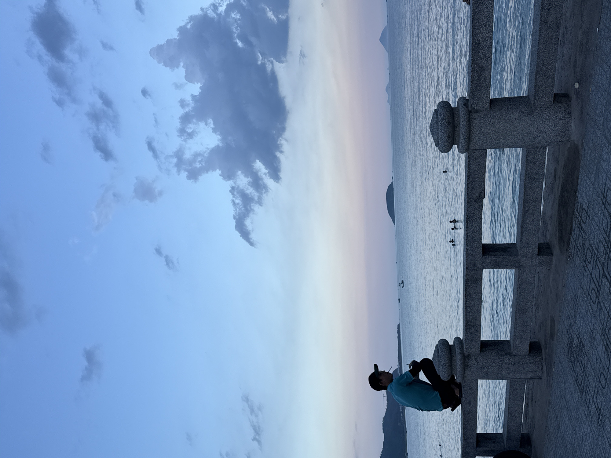 A person is sitting on a stone railing overlooking the sea. The person is wearing a blue shirt and a baseball cap. They are looking out at the water, with their legs crossed. The sea is calm, and there are a few small boats in the distance. The sky is a gradient of blue and pink, suggesting it is either sunrise or sunset. The railing is made of stone and has decorative carvings. The ground beneath the railing is paved with patterned tiles. The scene is peaceful and serene. The time of day is likely dusk or dawn, given the sky colors. The weather appears to be clear and calm. The location is a waterfront, possibly a promenade or pier, in Bac Nha Trang Ward, Vietnam. There are no visible text elements in the image.