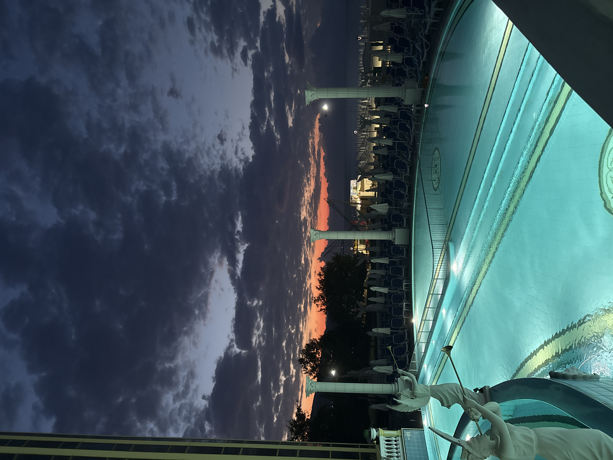 An outdoor swimming pool area is depicted at dusk, with the sky displaying shades of dark gray clouds tinged with orange and red near the horizon. The pool is illuminated with turquoise lighting, creating a vibrant glow. Two classical white statues of winged figures, appearing to be cherubs or angels holding trumpets, are positioned on the left edge of the pool. The area surrounding the pool is furnished with numerous blue lounge chairs and white umbrellas, suggesting a resort or hotel setting. In the background, trees are silhouetted against the twilight sky, and further beyond, a body of water, likely the sea, is visible. Architectural elements resembling classical columns are present near the pool. The scene captures a serene and atmospheric moment as day transitions into night in Nesebar, Bulgaria.