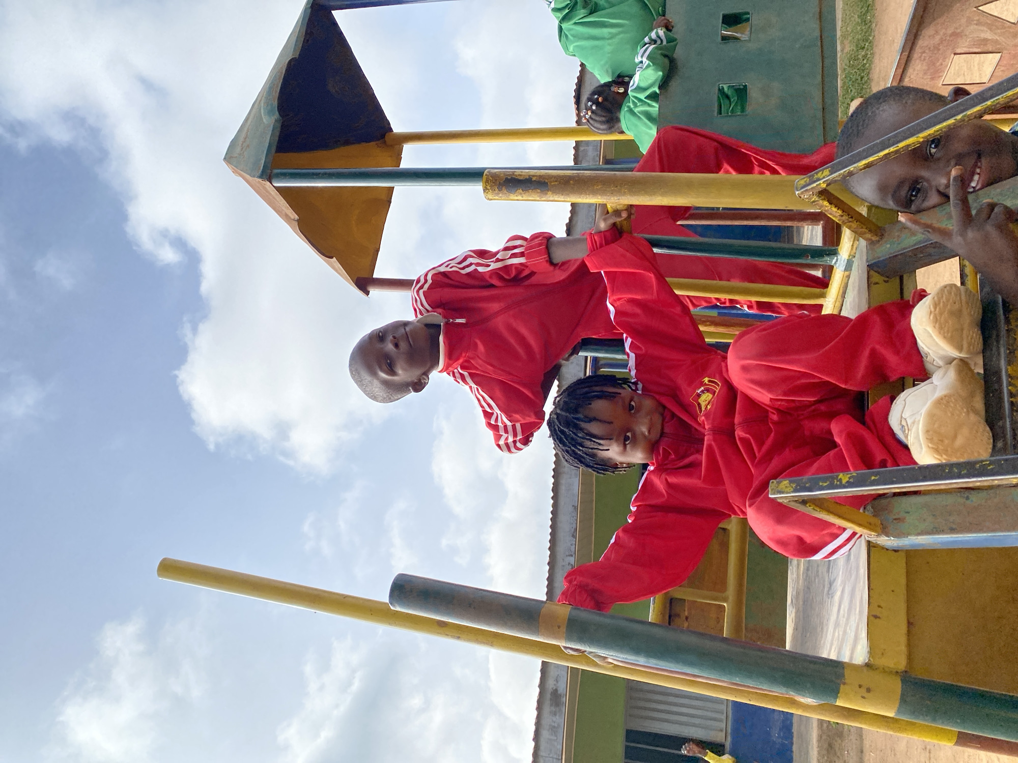 Captured in Gura Top, Nigeria, the image presents a vibrant playground scene. Three children are visible, two clad in matching red tracksuits with white stripes, while another wears a green tracksuit, all on a colorful metal playground structure. One of the children in red makes a "peace" sign towards the camera. The sky is partly cloudy. The playground equipment appears aged with visible wear and paint discoloration.