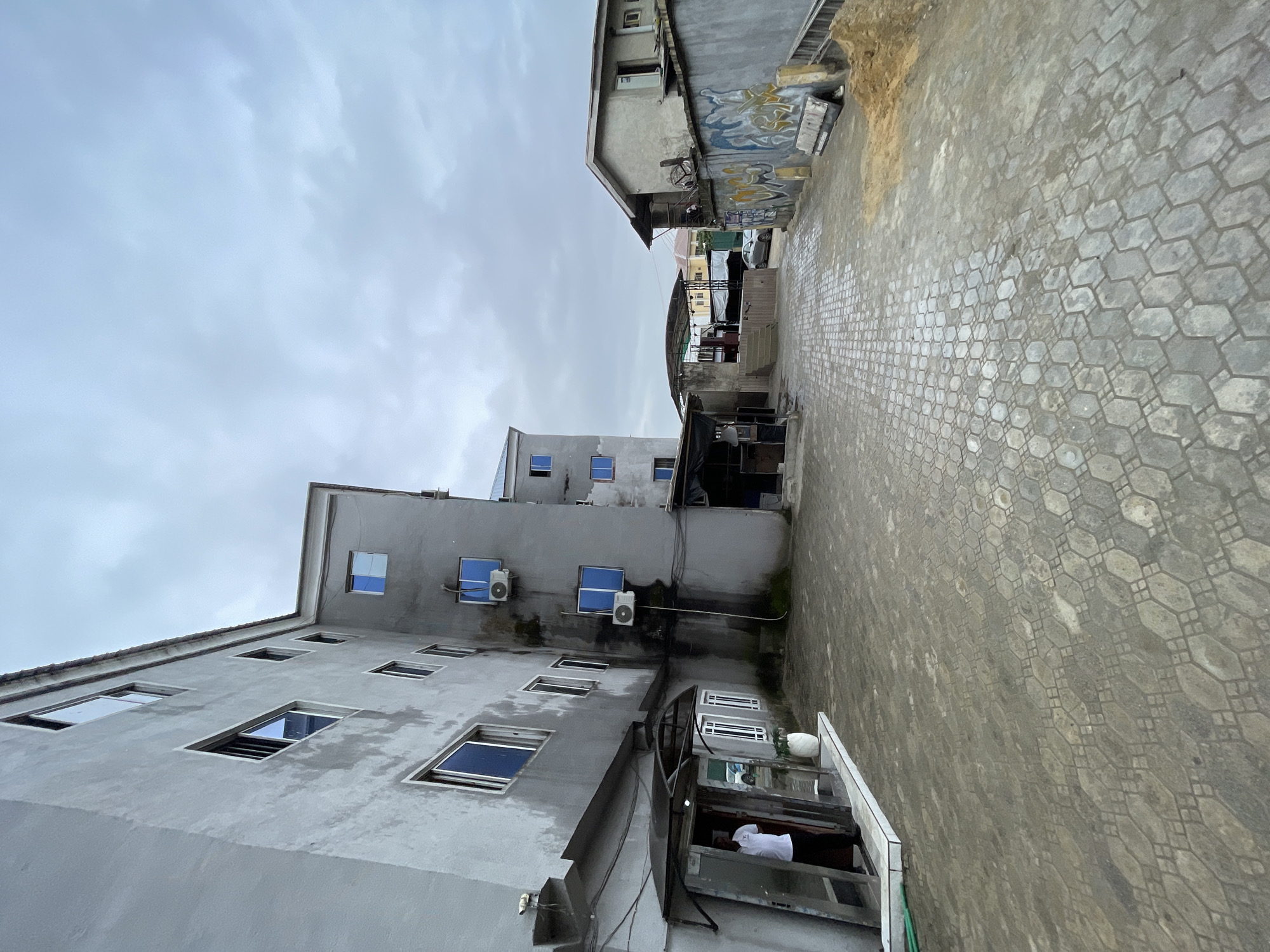 The image depicts an outdoor courtyard in Ado, Nigeria, with overcast skies. On the left, a multi-story building with a gray facade dominates the scene. The building has numerous windows with blue panes and visible air conditioning units mounted on its exterior. An open entranceway with a glass door is visible, and a person in a white shirt and dark pants stands just inside. A white decorative pot is placed near the entrance. The ground of the courtyard is paved with hexagonal interlocking concrete blocks. To the right, a single-story building with a similar gray facade is present, featuring a sloped roof and a wall covered in colorful graffiti. Adjacent to this building is a pile of sand and construction debris, with a ladder leaning against the wall. Further in the background, additional buildings and a covered structure with seating are visible. A silver car is parked on the paved area. The overall impression is an urban environment with signs of ongoing construction or recent activity.