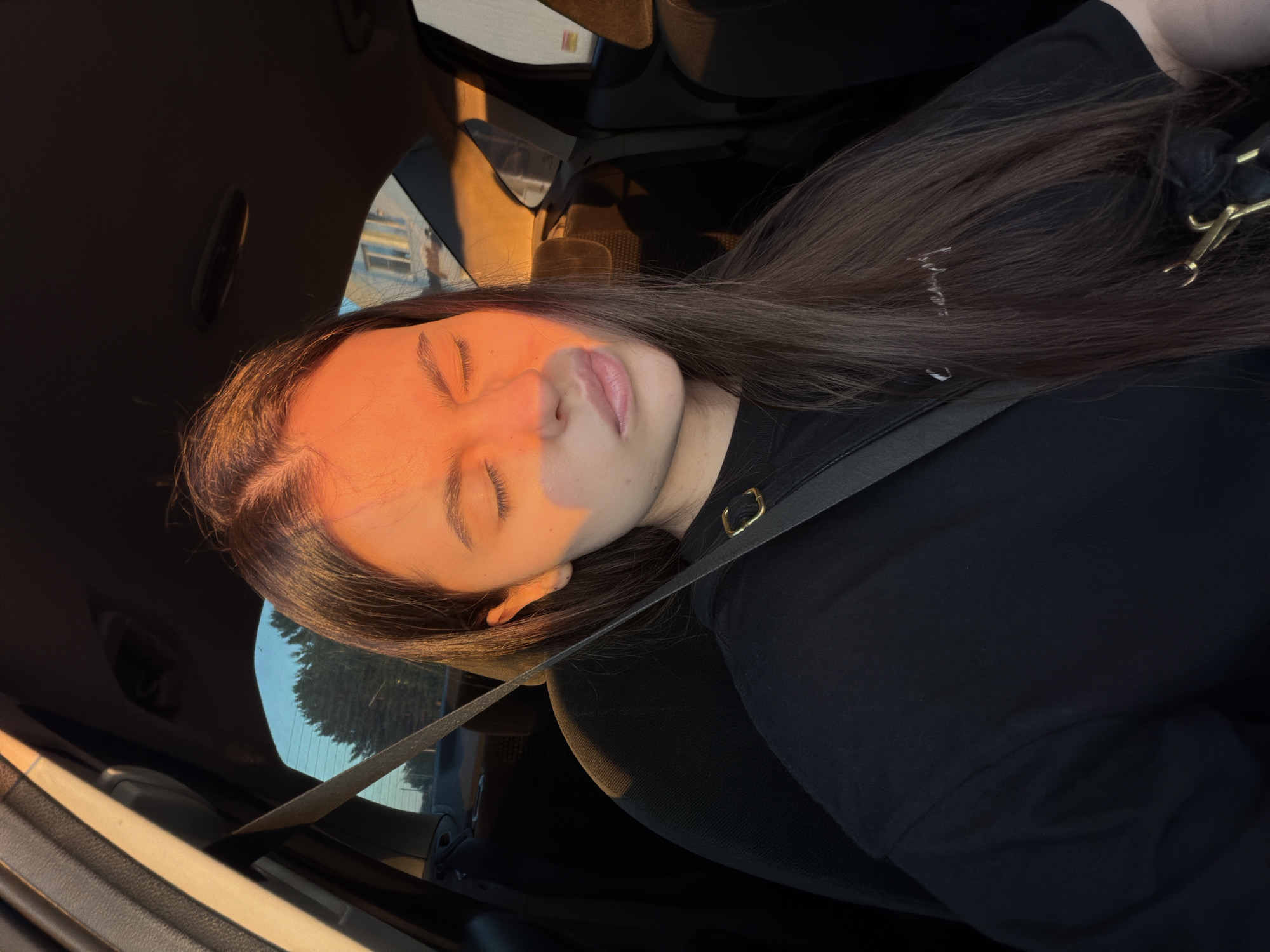 The image shows a young woman with her eyes closed, sitting in the passenger seat of a car. Sunlight casts an orange glow across her face and hair. She is wearing a black t-shirt and a seatbelt is visible across her chest. The interior of the car, including the dashboard and seats, is dark. Outside the car window, trees and buildings are visible under a blue sky, indicating an outdoor setting, likely during the daytime. The specific location in the city of Chaikovsky, Russia, cannot be determined from the visual information alone.
