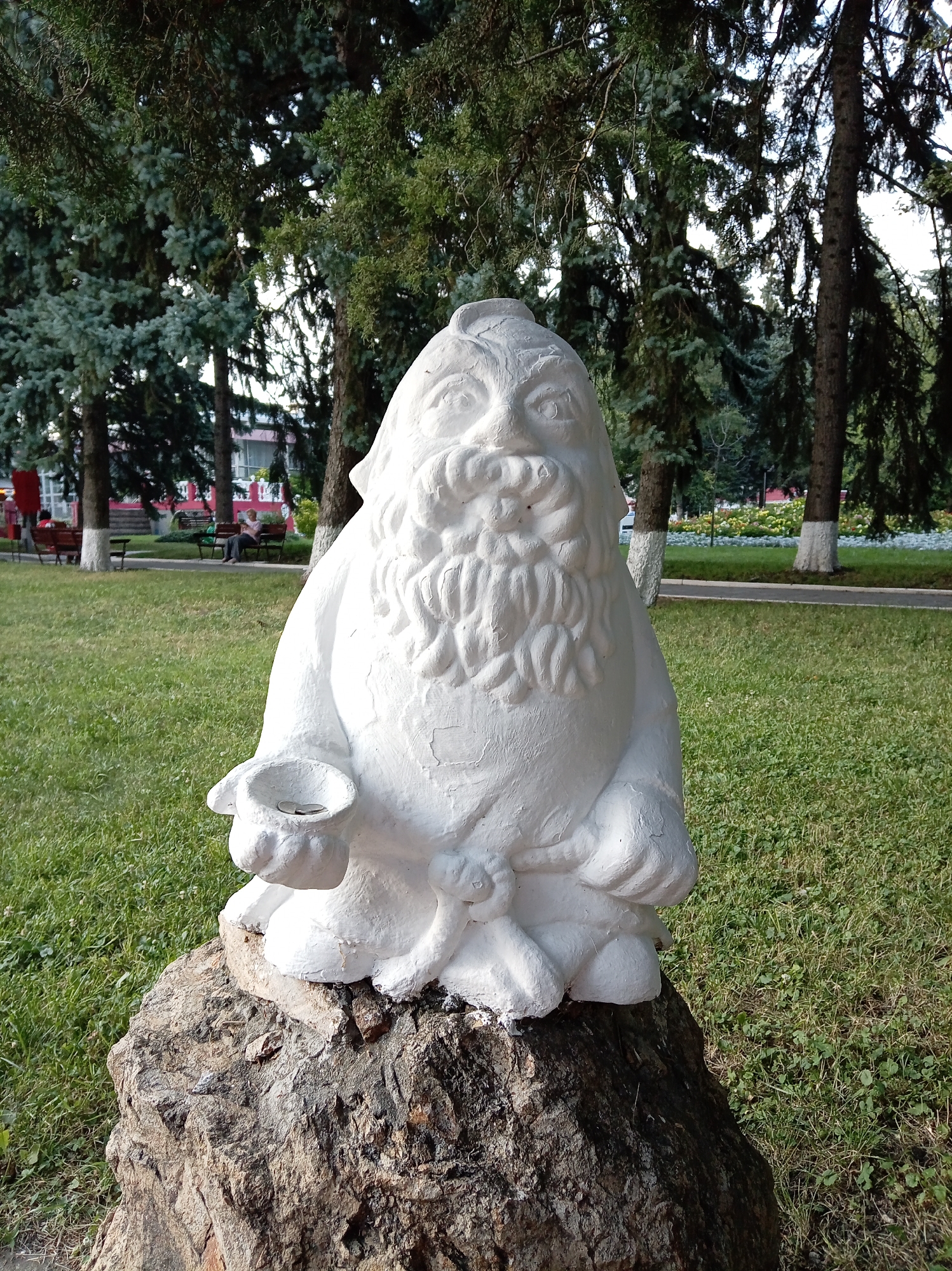 In Yessentuki, Russia, a white, stylized sculpture of a bearded gnome sits atop a large, rough-hewn rock in a grassy park area. The gnome is depicted with a round body, a prominent beard, and is holding out a cupped hand containing two coins. Behind the sculpture, trees with lush green foliage are visible, along with park benches and people in the background. The scene appears to be an outdoor public space, likely a park or garden within the city.