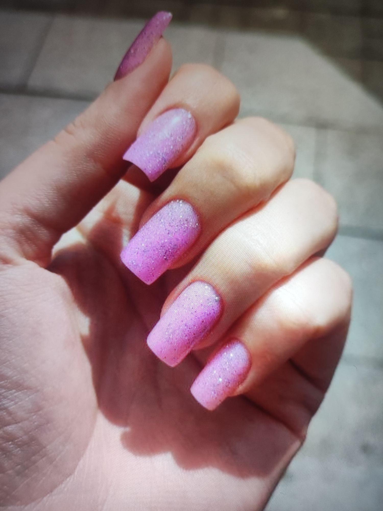 A close-up shot focuses on a person's hand with several nails adorned with pink glitter. The background appears to be an outdoor setting, possibly a sidewalk or paved area, with some suggestion of a wall or building. The lighting indicates it's daytime. There is no information available to determine the location of this scene in the city of Cherkasy, Ukraine.