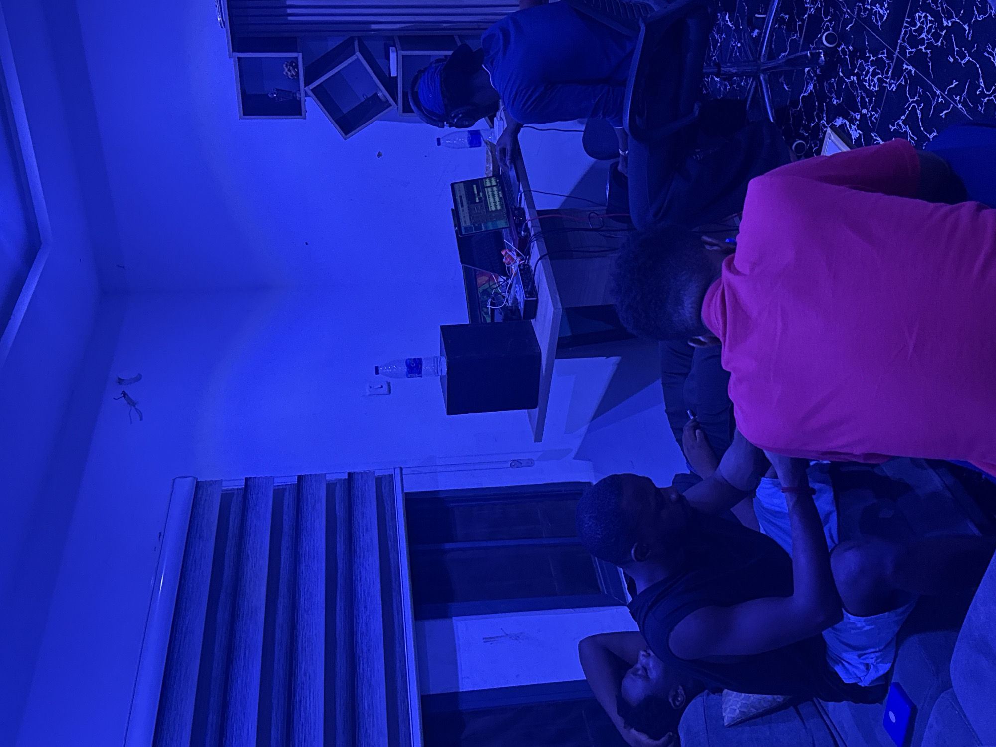 The image shows an indoor scene with three individuals, possibly in a recording studio or creative space. The individuals are all African men. One man is seated at a desk, wearing headphones and working on a computer with audio editing software visible on the screen. He is wearing a blue shirt and a bandana. Another man is standing next to him, wearing a pink shirt. A third man is lounging on a couch in the foreground. The room is lit with blue lighting, and there are water bottles, speakers, and other equipment scattered around. The location is likely a room in Abuja, Nigeria, based on the demographics of the people and the general setting. The walls are painted white, and one wall features a black and white decorative pattern. Window blinds are visible.