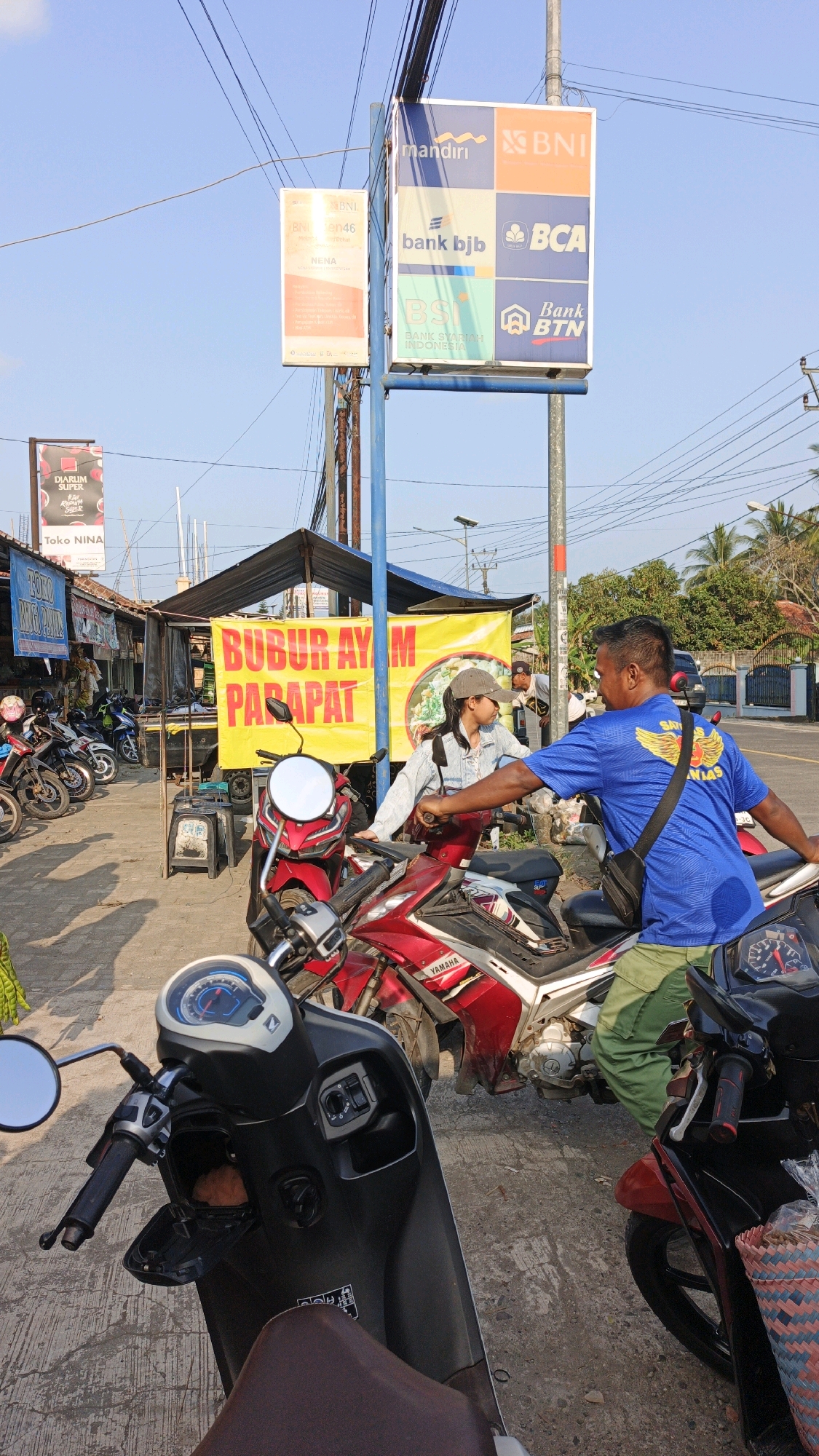 A street scene in Cibenda, Indonesia, features a food stall with a large yellow banner that reads "BUBUR AYAM PARAPAT" in red lettering. A woman wearing a gray cap and denim jacket is standing next to a red motorcycle, interacting with a man in a blue t-shirt and green pants who is seated on another motorcycle. Several other motorcycles are parked in the background. Above the stall, a blue pole holds signs for various Indonesian banks, including Mandiri, BNI, bank bjb, BCA, BSI, and Bank BTN. The sky is clear and blue, and the ground is paved.