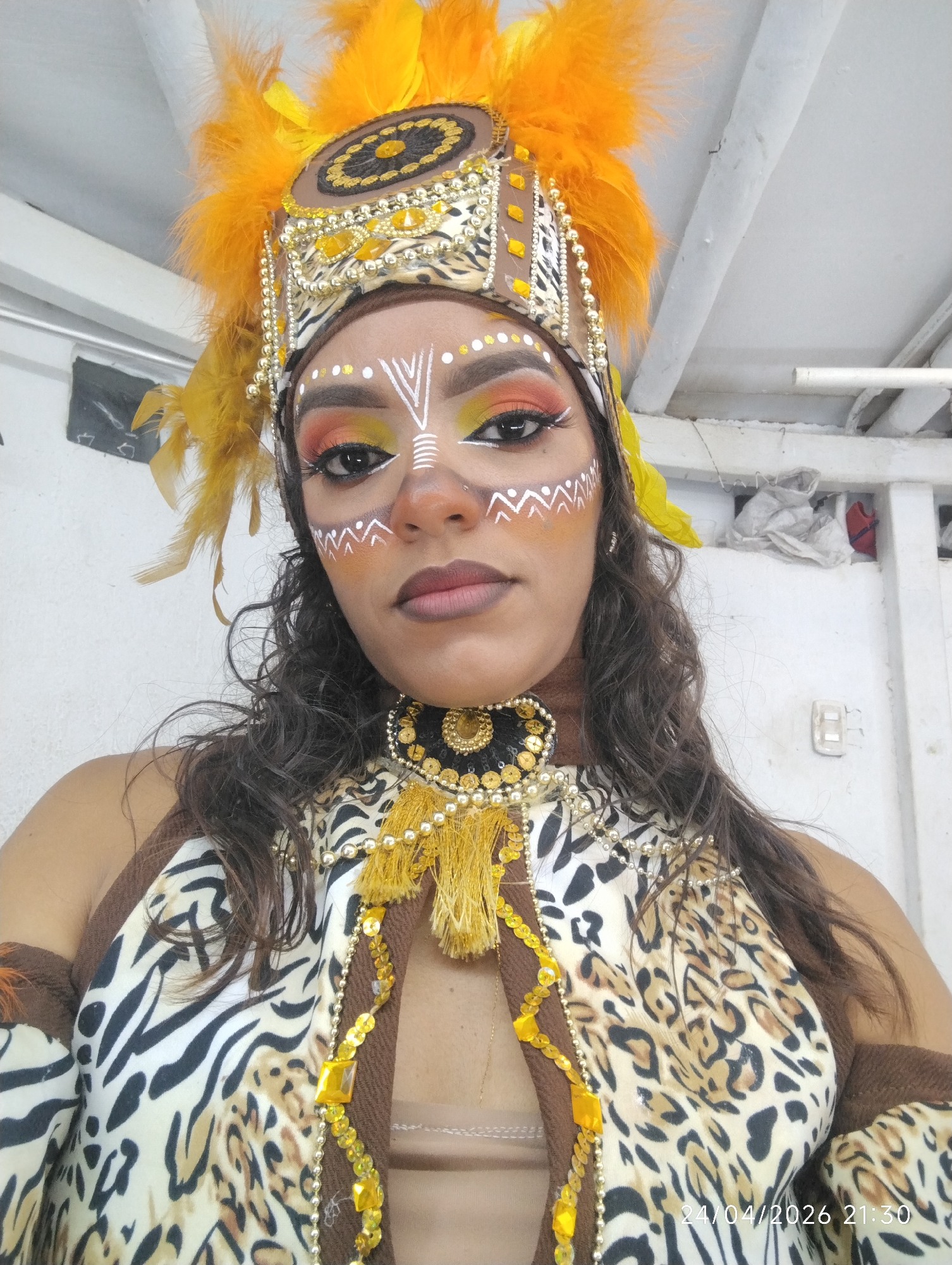 The image shows a woman in tribal-inspired attire, including a headdress with orange feathers, face paint with white markings, and a leopard and zebra print top with yellow embellishments. She is wearing a necklace with a black and gold pendant. The background is a plain, white wall. The picture was taken on 24/04/2026 at 21:30 in an unknown city.