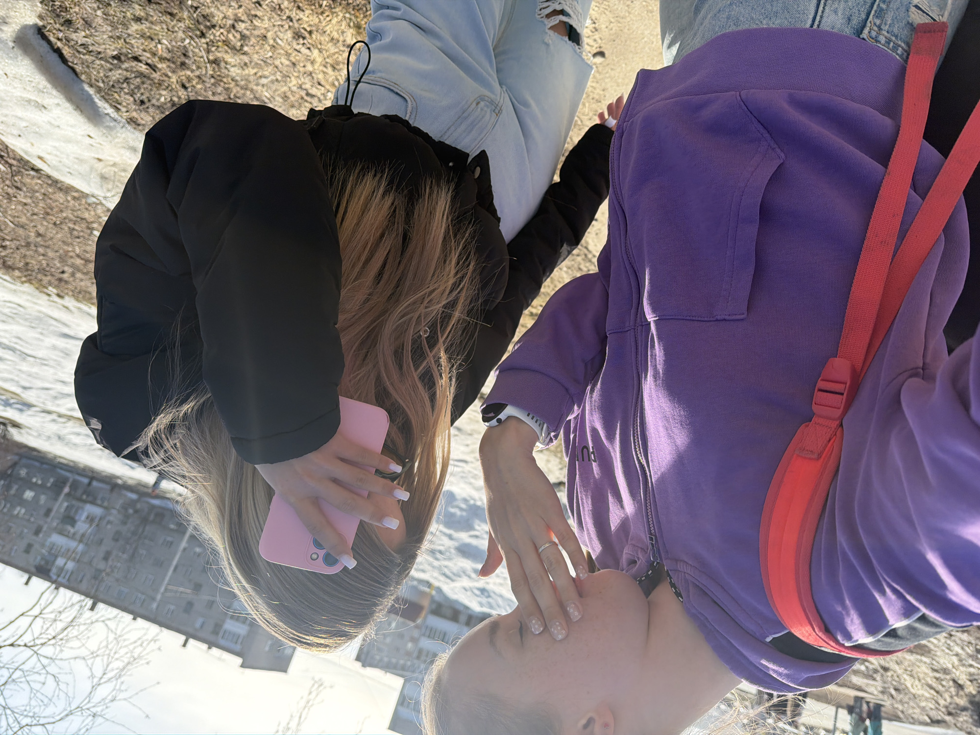 Two women stand outdoors. The woman on the left wears a black puffer jacket and light-wash ripped jeans, and she holds a pink cell phone. The woman on the right wears a purple zip-up hoodie and has a red strap across her body. They are standing in what appears to be a city park with bare trees and melting snow. A large apartment building is visible in the background, indicating a city setting. Based on the information provided, this location is in Berezniki, Russia.