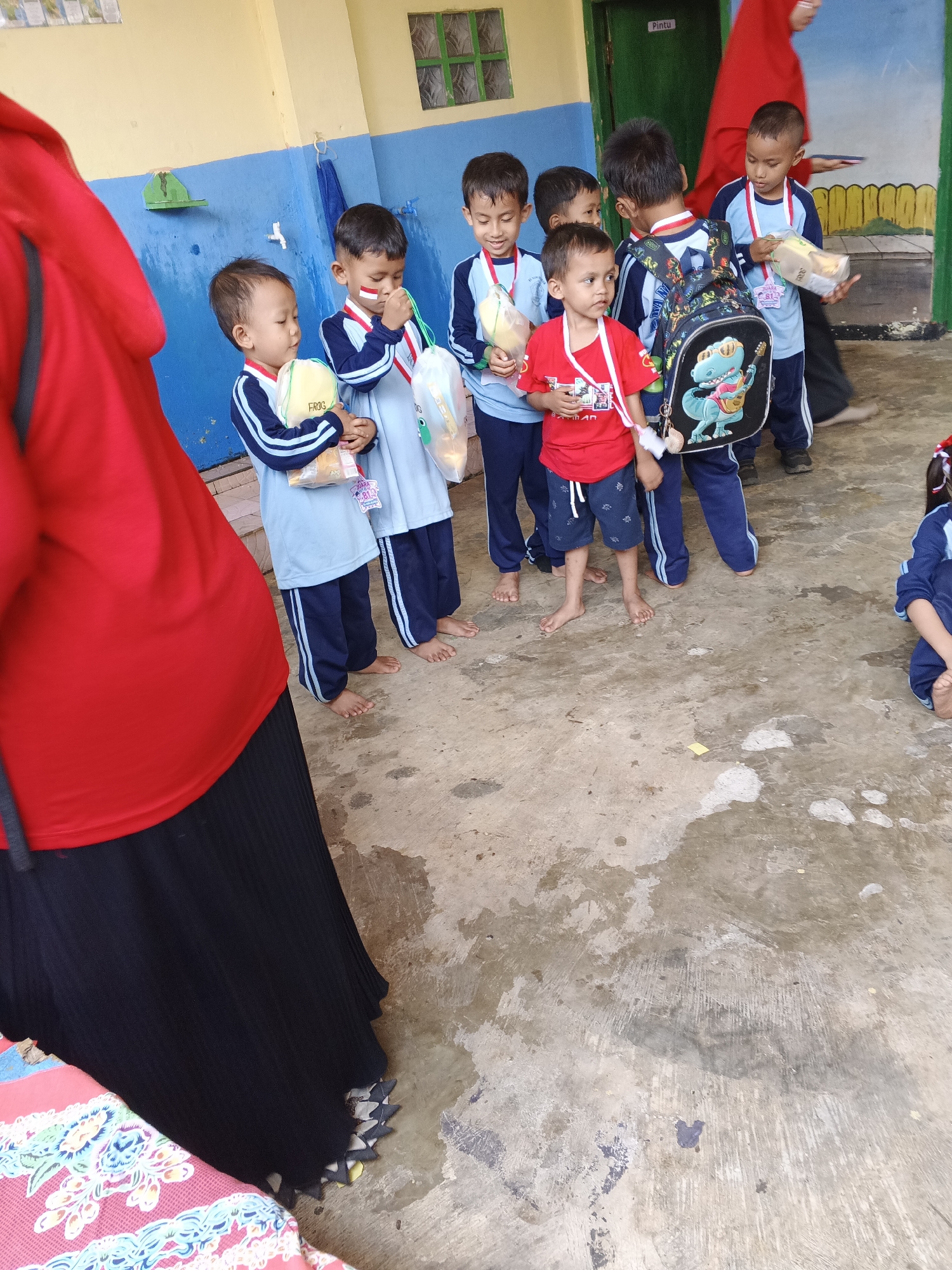 A group of young children, likely students, stand on a concrete ground in front of a yellow and blue painted building. The children are wearing light blue uniforms with dark blue accents and some have medals around their necks. They are holding various items, including bags of snacks and small toys. One child wears a red t-shirt and blue shorts. A person in a bright red hijab and black skirt stands to the left, partially obscuring the view of some of the children. The environment appears to be an outdoor school area, with a green-painted shelf and a blue towel hanging on the wall of the building. The ground is wet in places, suggesting it may have recently rained. The overall scene depicts a casual gathering of children, possibly during a school event or break time. The location is identified as Warungkiara, Indonesia.