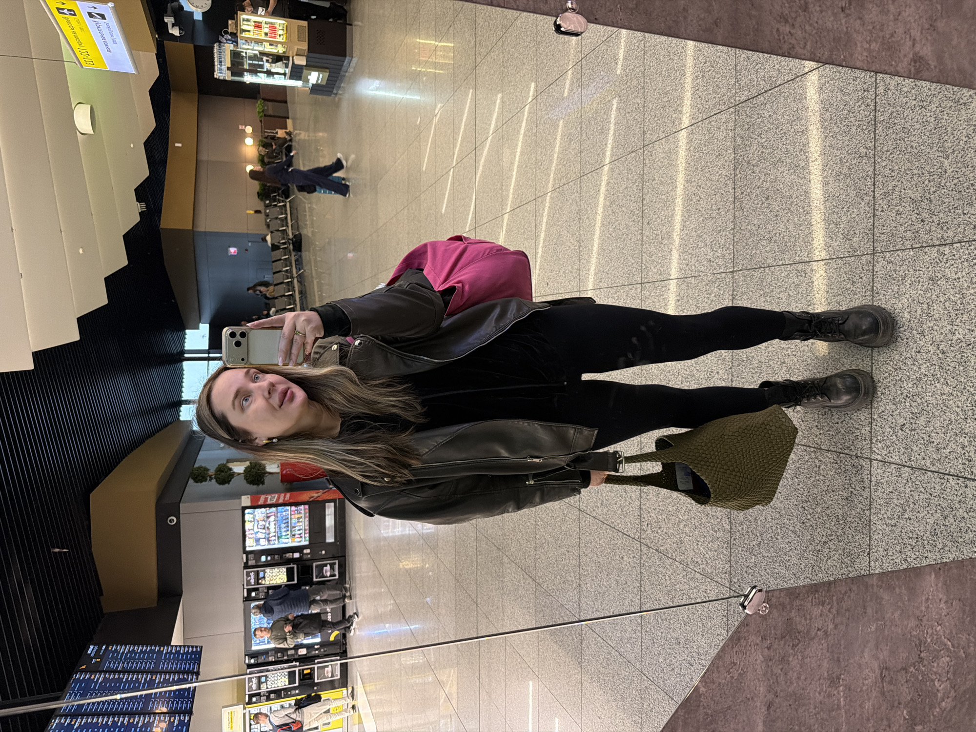 A woman with long blonde hair is taking a selfie in an airport-like setting. She is wearing black leggings, a black top, a black leather jacket, and black boots. She is holding a green textured bag in her left hand and a smartphone in her right hand. She has a pink backpack. The background includes airport seating, vending machines, and flight information displays. The woman appears to be standing in a reflection, likely on a polished floor or wall. Based on the appearance of the environment, it may be the Sheremetyevo International Airport in Khimki, Russia.