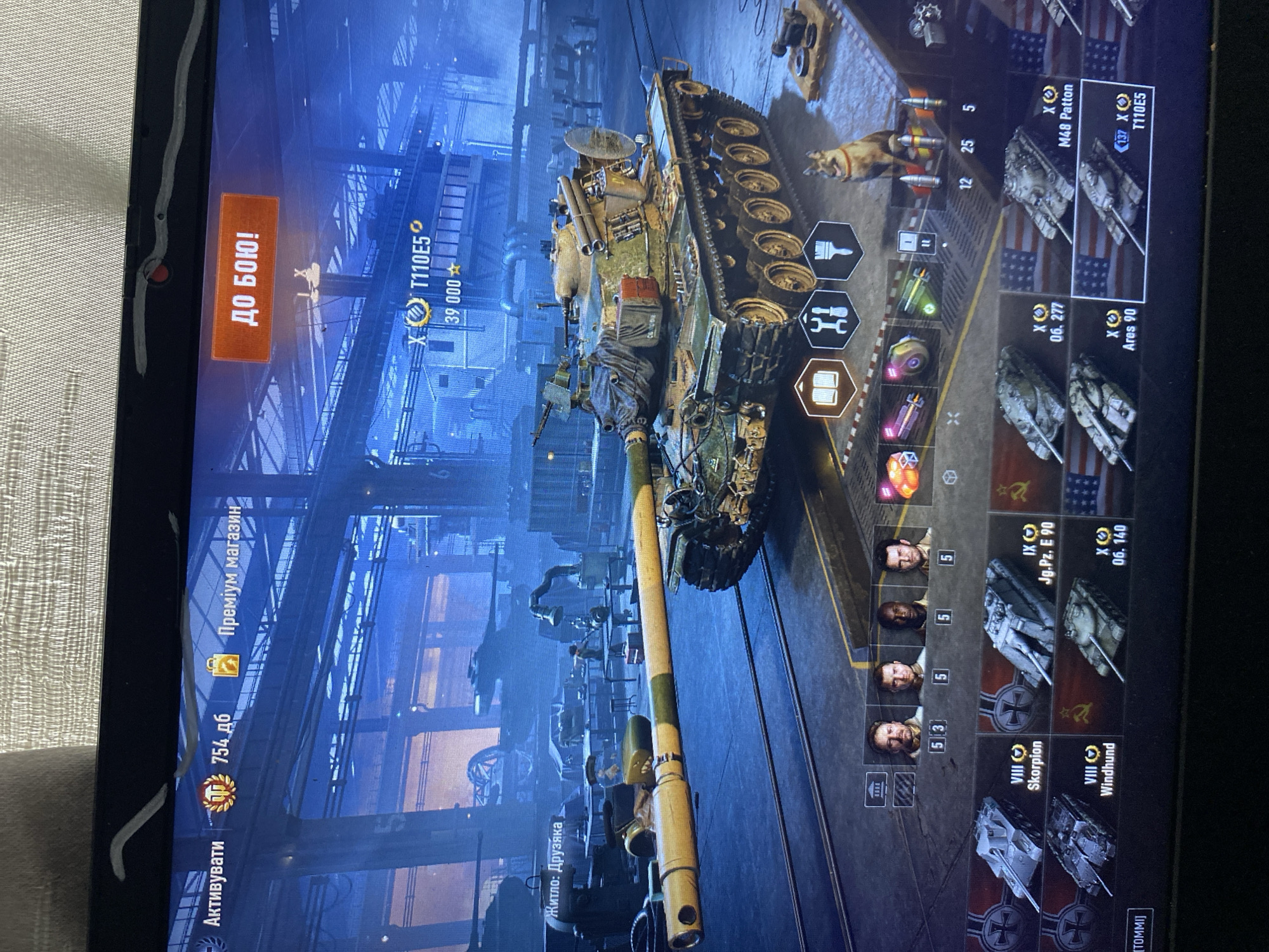 The image depicts a screenshot from a video game, likely World of Tanks, displayed on a laptop screen. The game interface shows a military hangar environment with various tanks. In the foreground, a green and yellow tank, identified as T110E5, is prominently displayed. The upper portion of the screen features game-related text and buttons, including "Активатувати" (Activate), "754 дб" (754 db), "Преміум магазин" (Premium Shop), and a large orange button that reads "ДО БОЮ!" (To Battle!). A small dog is visible to the right of the tank. Below the main tank, a carousel displays several other tank models with their names and tiers, such as "VIII Scorpion," "VIII Windhund," "IX Jg.Pz. E 90," "X 06. 277," "X Ares 90," "X M48 Patton," and "T110E5." The image does not contain any information or visual cues that would place it in the city of Szczecin, Poland.