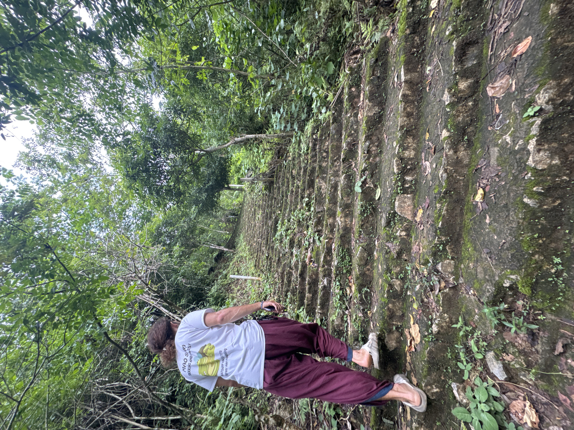 A person with long, curly brown hair, wearing a white t-shirt with a graphic and text, and loose-fitting maroon pants, walks up a moss-covered stone staircase in a lush, green forest. The staircase appears old and weathered, with vegetation growing between and around the stones. The surrounding environment is dense with trees and foliage, indicating a natural, possibly remote, setting. The scene is set in Mai Chau, Vietnam, suggesting a trekking or sightseeing activity.