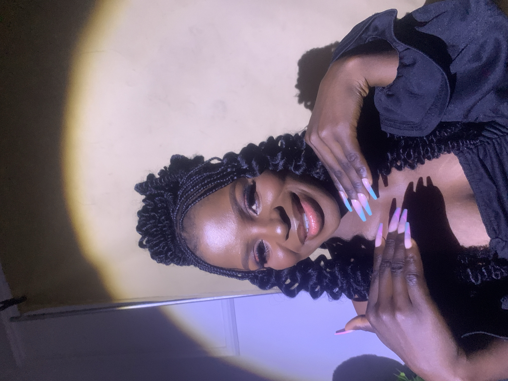 A young Black woman with braided hair is posing indoors. She is wearing a dark top with frills and has long, pastel-colored manicured nails. She is smiling. The lighting is uneven, with a bright spot on the wall behind her. The location is likely Jos, Nigeria.