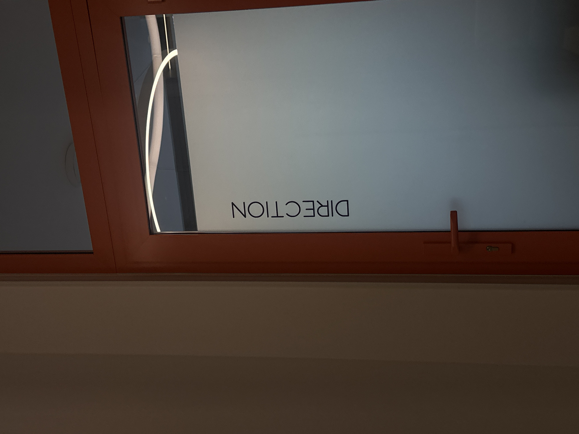 A window with the word "DIRECTION" printed backwards on it, likely to be read correctly from the other side. The window is framed with a reddish-brown painted wood. The wall below the window is a light tan color. The image was taken inside. It is not possible to determine the specific location within Paris from the given image.