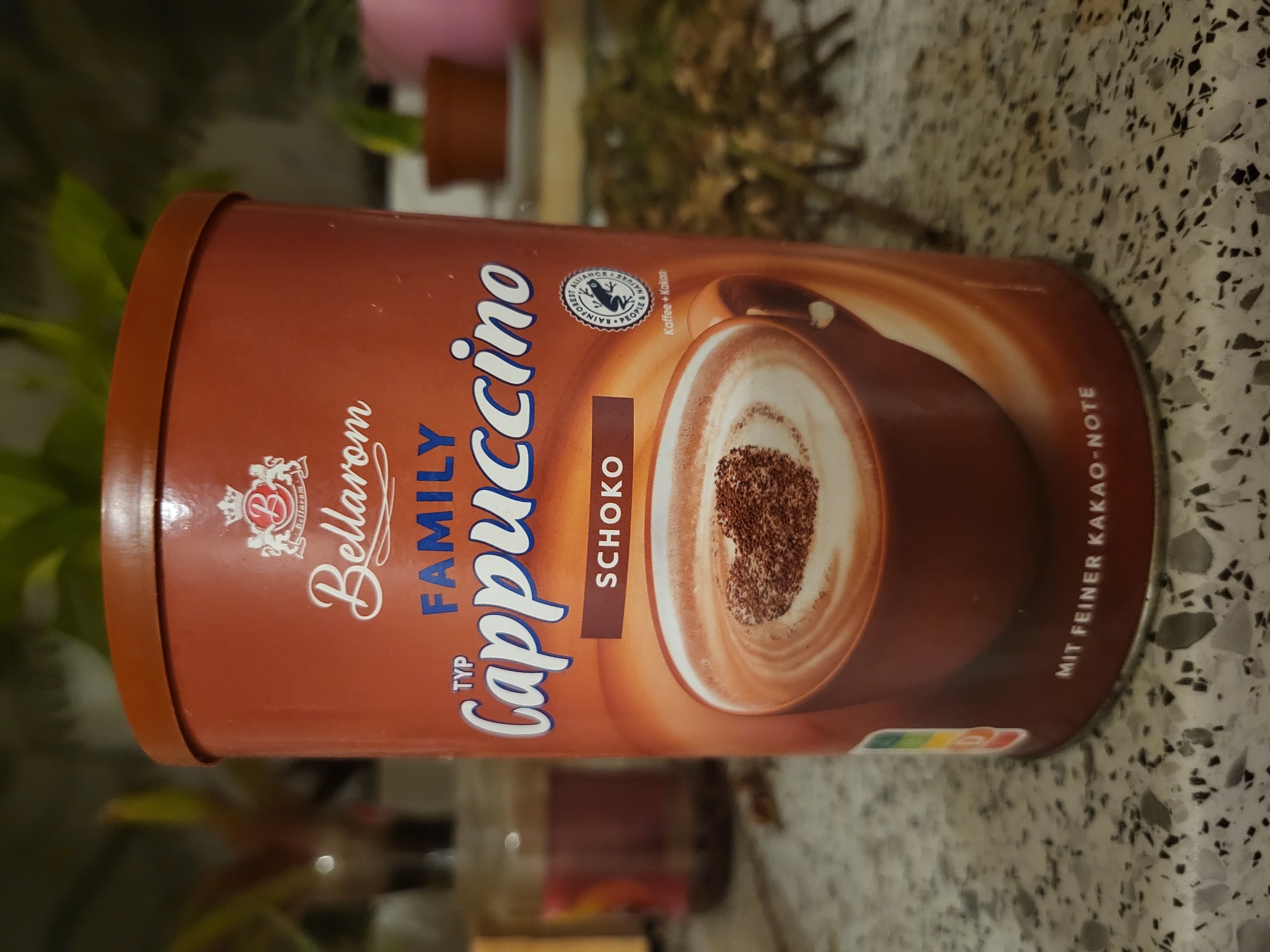 A brown tin of Bellarom Family Cappuccino is displayed on a speckled countertop. The tin features the Bellarom logo, the words "FAMILY CAPPUCCINO," "SCHOKO," and "MIT FEINER KAKAO-NOTE." A drawing of a cup of cappuccino with a heart-shaped sprinkle of cocoa is prominently featured. A Rainforest Alliance seal is visible on the right side of the tin. The background shows out-of-focus plants and a jar. This scene is located in the city of Pinneberg, Germany.