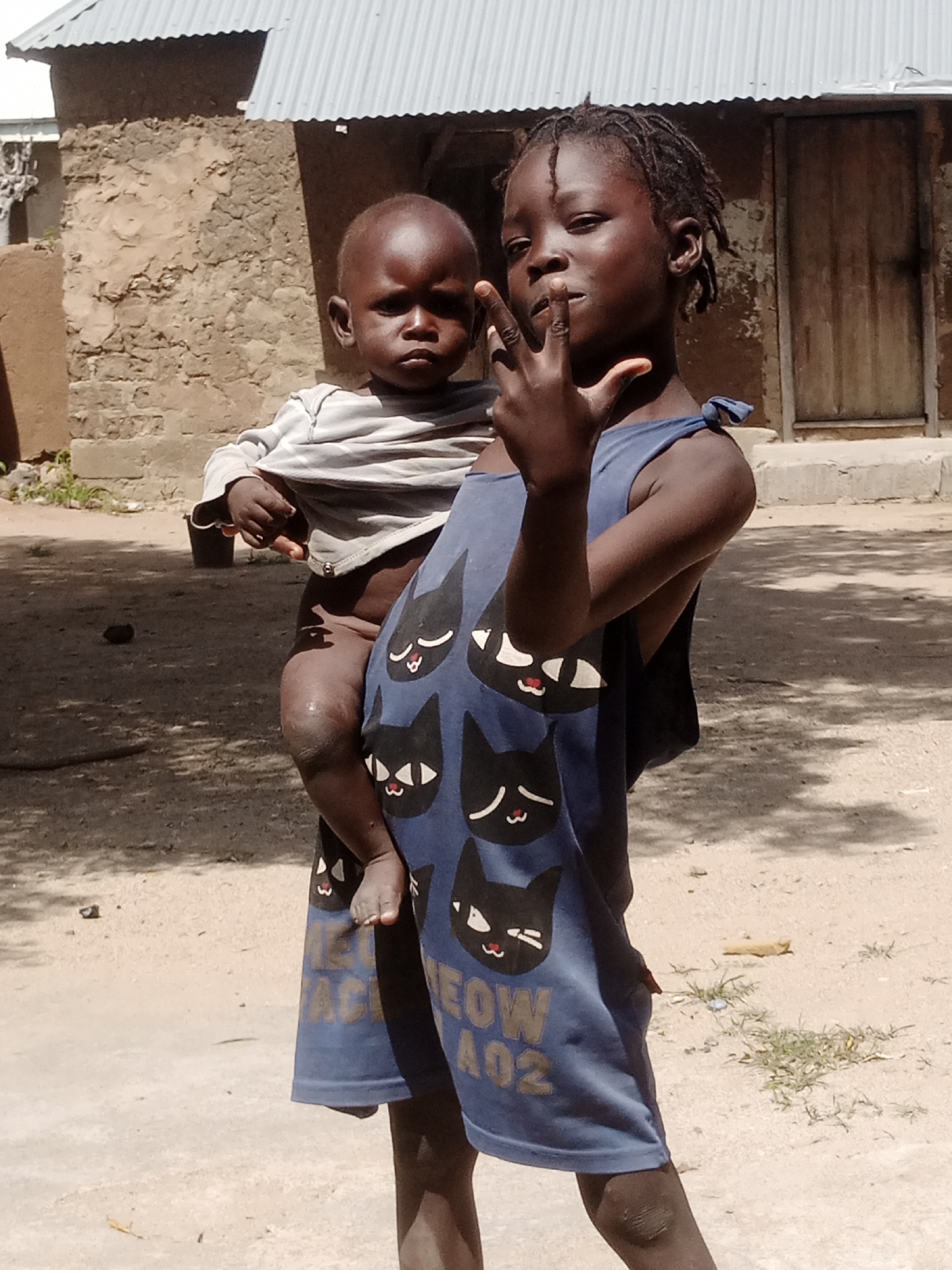 In Langtang, Nigeria, two children stand in a dusty, outdoor environment. The older child, on the right, wears a blue sleeveless dress adorned with cartoon cat faces. This child is holding a younger child, who is partially visible and dressed in a grey and white striped shirt. The older child's right hand is raised, with their index and middle fingers extended, suggesting they are gesturing or posing. In the background, a simple building with a corrugated metal roof and adobe walls is visible. The ground is sandy and uneven, with patches of dry grass. The lighting suggests a sunny day.