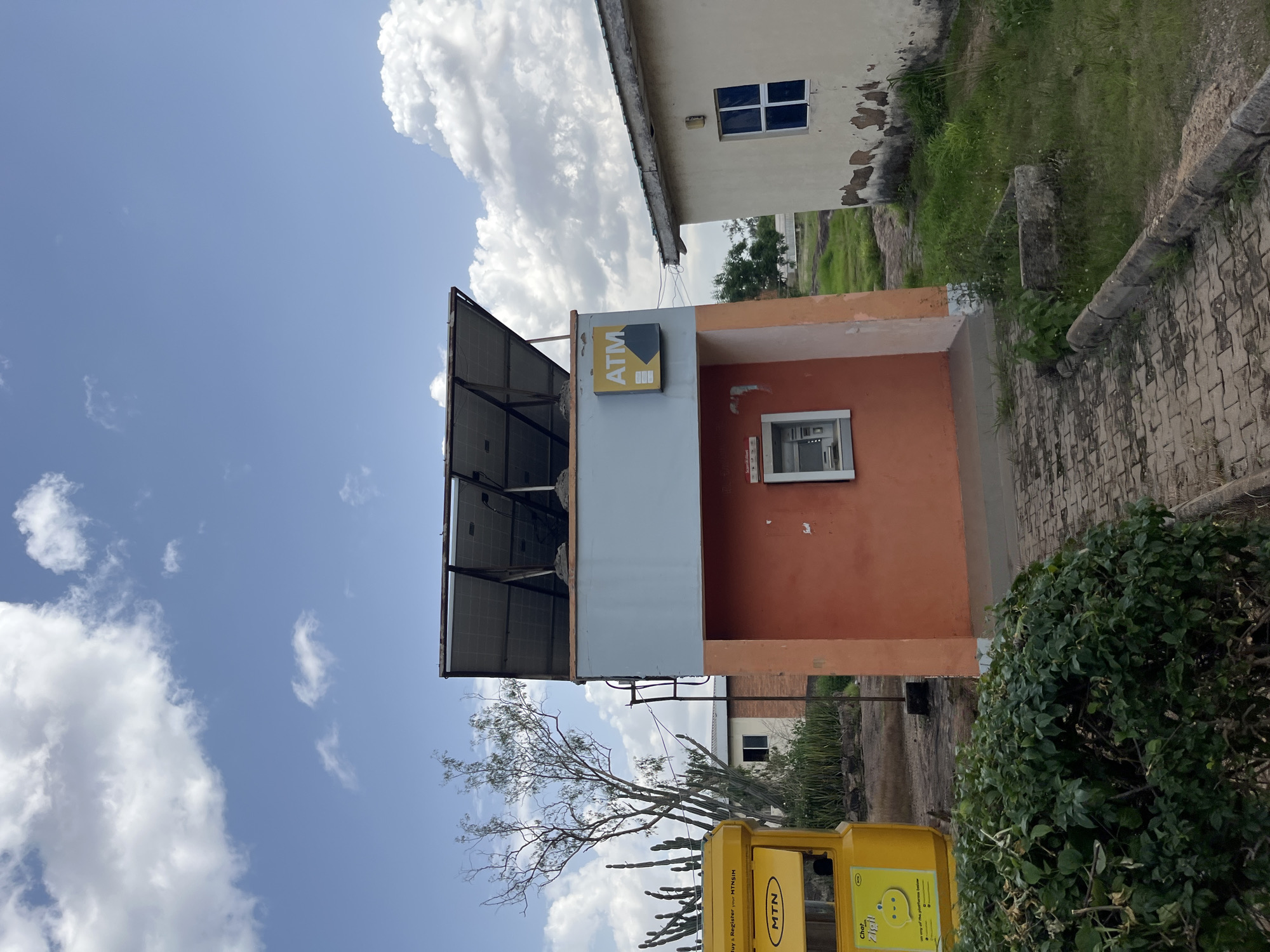 An outdoor, ground-level view captures an ATM kiosk in Jos, Nigeria, under a partly cloudy blue sky. The kiosk, painted orange on its lower half and light blue on its upper half, features an ATM machine embedded in its front. Above the ATM, a yellow sign with "ATM" in black lettering and a gray arrow is affixed to the blue section. Solar panels are mounted on the angled roof of the kiosk. To the left, a yellow MTN-branded structure, possibly a mobile money booth, is visible with some text and a graphic on its side. In the background, there are buildings, some with visible signs of wear, and various types of vegetation, including cacti and trees. A paved walkway leads towards the ATM kiosk, bordered by grass and a low concrete curb.