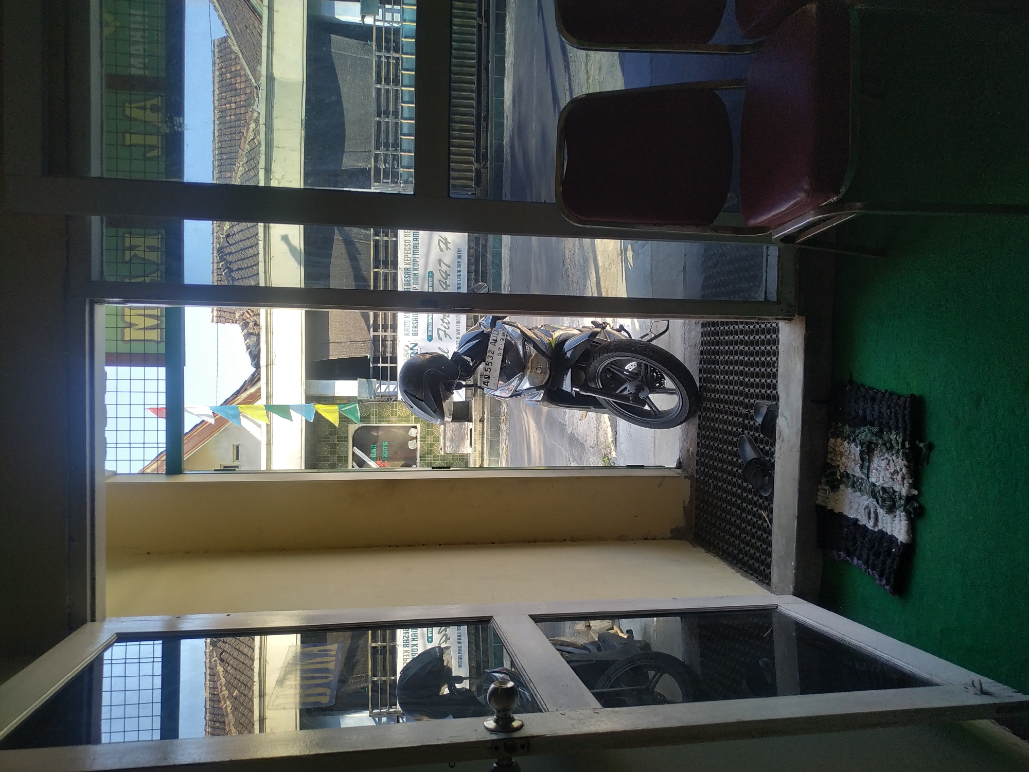 This is a medium-angle, full-shot photograph taken during the daytime, looking outwards from an interior space with a green carpeted floor through a glass doorway. The doorway frame is light beige on the left and a darker metallic color on the right. To the left of the doorway, a glass door is open, revealing a reflection of a black motorcycle. Inside the doorway, a black motorcycle with a helmet on its handlebars is parked. The motorcycle has a license plate with the characters "AQ 5532 AUD". Further inside, two red chairs are visible. Outside the doorway, a street is visible with buildings and tiled roofs. A string of triangular flags in various colors, including yellow, blue, and red, hangs across the street. A portion of a sign with text that reads "M A JAL" is visible on a building. In the foreground, a small, multi-colored rag rug lies on the green carpet. A pair of black flip-flops is placed on a black mat near the doorway. The overall scene suggests an entrance to a business or residence in a city. While the location is stated as Batan, Indonesia, the image itself does not contain any specific visual cues to definitively confirm this location.
