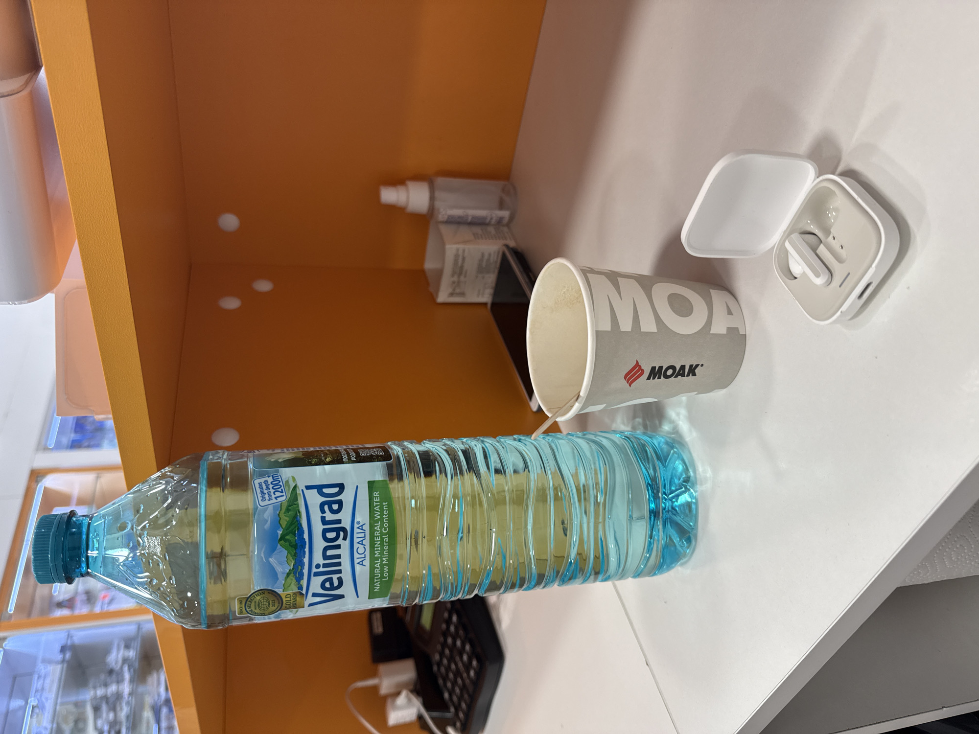 The image shows a still life arrangement on a white countertop, likely indoors. The objects present include a bottle of "Velingrad Alcalia" natural mineral water, a disposable coffee cup labeled "MOA," a pair of white wireless earbuds with their charging case, a small spray bottle, a box and a black keyboard, and some assorted charging cables. The water bottle is positioned horizontally, with its label prominently displayed. The coffee cup sits nearby, next to the earbud case. The countertop appears to be against an orange shelf. The location of Burgas, Bulgaria is not explicit but can be inferred from the "Velingrad" branded water.