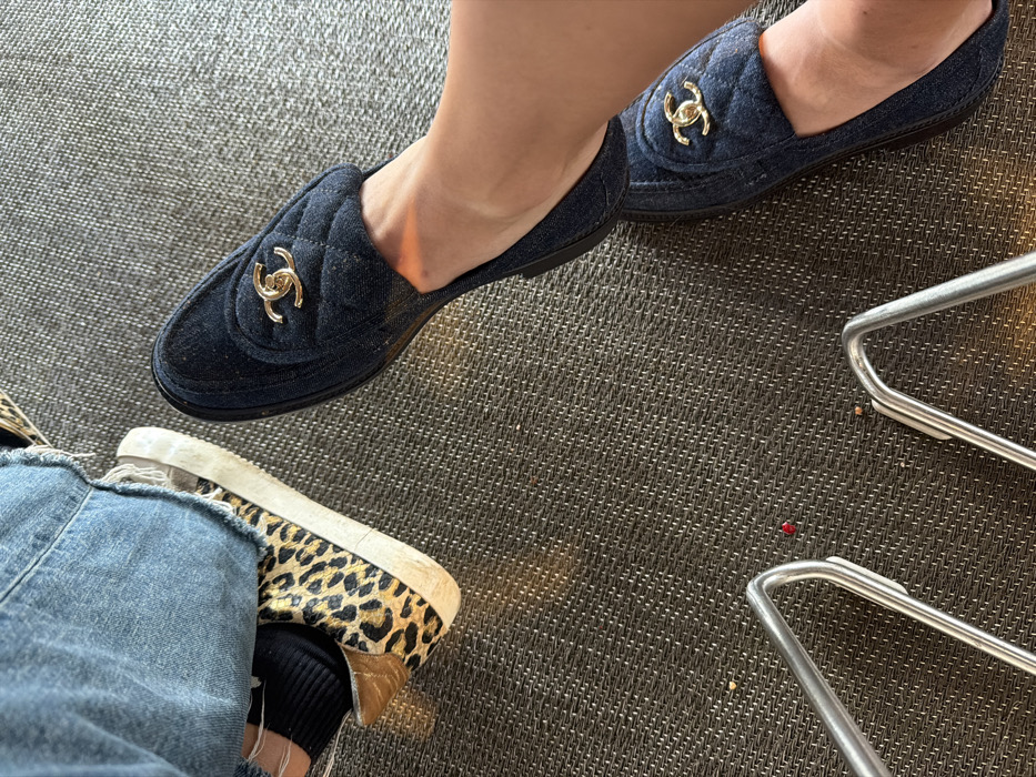 The image captures a close-up, high-angle view of two sets of feet, likely belonging to two different individuals, resting on a textured, dark gray or brown carpeted floor. The overall impression is one of a casual indoor setting, possibly a waiting area or a lounge.
In the upper half of the image, a person's bare foot and lower leg are visible, wearing a dark blue, possibly denim, quilted slip-on shoe. This shoe features a prominent gold-colored interlocking "CC" logo, indicative of the Chanel brand, positioned on the vamp. The shoe appears to be a flat or mule style. Only one foot and shoe are clearly visible, with a glimpse of the second matching shoe further back on the right side. The skin tone of the leg is light.
In the lower left quadrant of the image, the leg and foot of another person are visible. This person is wearing distressed light blue denim jeans, characterized by ripped details around the knee area. A black ribbed sock is visible below the jeans. The foot is clad in a casual shoe, possibly a sneaker or slip-on, featuring a distinctive leopard print pattern on the upper part, made from what appears to be a textured fabric like faux fur or pony hair. The sole of this shoe is white, showing some signs of wear and dirt. A small part of a shiny, possibly metallic, gold-colored detail is visible on the heel area of this shoe.
The floor is covered with a tightly woven, dark-colored carpet or mat, exhibiting a subtle geometric or basket-weave texture. In the bottom right corner, part of a metallic, silver-colored chair leg or table frame is visible, consisting of a rounded rectangular shape. On the carpet near this metallic frame, a small, bright red object, possibly a piece of food or debris, is noticeable, along with a few other tiny light-colored specks.
The lighting in the image suggests an indoor environment, with soft shadows and a slightly warm tone, particularly on the skin of the foot wearing the Chanel shoe. No text is explicitly visible beyond the brand logo on the shoe.