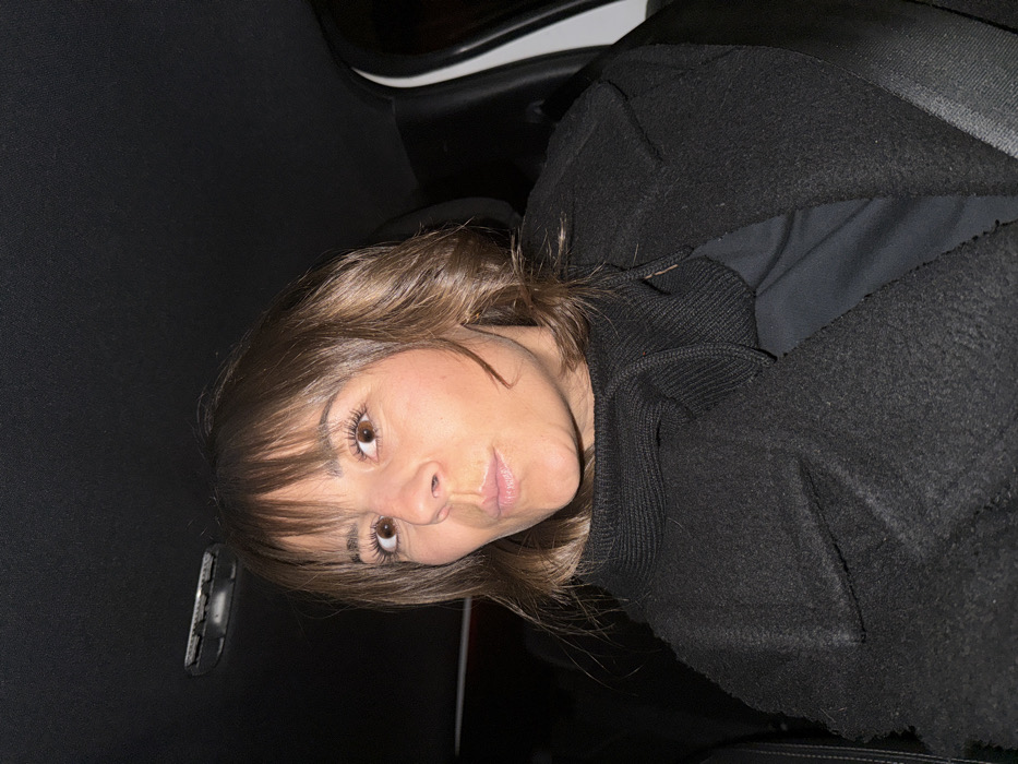 A woman with brown hair and bangs is looking at the camera with a slightly quizzical expression. She's wearing a dark, possibly black, hooded sweatshirt or jacket. The background appears to be the interior of a car, given the dark upholstery. The lighting is subdued, but her face is illuminated enough to discern her features. The image is rotated slightly.