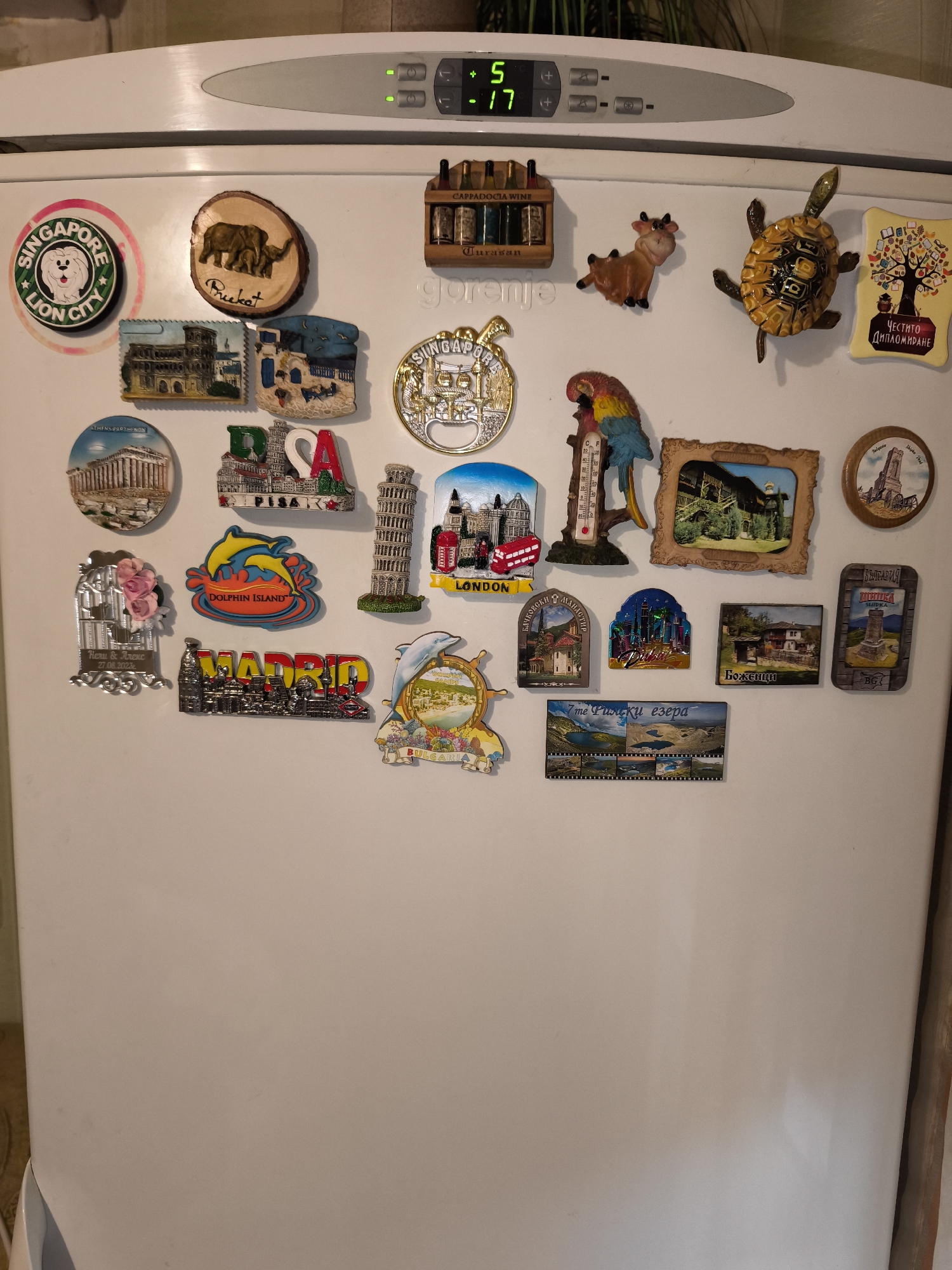 The image shows a white refrigerator door covered with various souvenir magnets. These magnets represent different locations and themes, including Singapore (Lion City), Phuket, Cappadocia Wine, Athens Parthenon, Pisa, Dolphin Island, Madrid, London, the Rila Lakes in Bulgaria, and individual Bulgarian cities. A few magnets show animals, like a cow and a turtle. There is also a parrot-shaped thermometer. The refrigerator's display shows a temperature reading of 5 and -17. The image doesn't contain any indication of Burgas, Bulgaria.