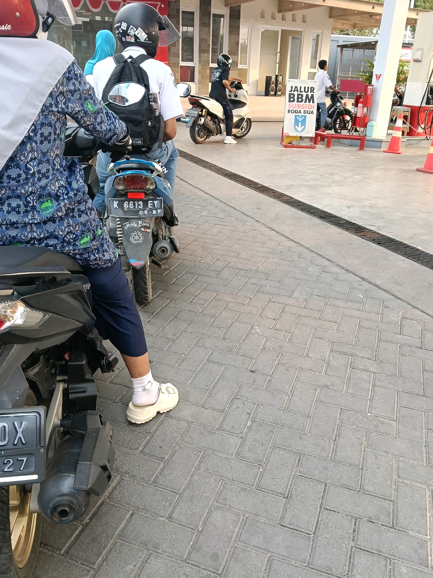 The image depicts a scene at a gas station in Tambakromo, Indonesia. In the foreground, a person wearing a blue batik-patterned shirt, navy blue capri pants, white socks, and cream-colored platform shoes is seated on a black motorcycle. The motorcycle has a license plate that reads "K 6613 EY". Behind this motorcycle, another motorcycle is visible with a rider wearing a black helmet and a backpack. Further in the background, two more individuals are on motorcycles, one wearing a blue headscarf and the other a white shirt. A sign in the center of the image reads "JALUR BBM SUBSIDI RODA DUA" (BBM SUBSIZED LANE TWO-WHEELED VEHICLES). Orange traffic cones are present, indicating a designated lane for motorcycles. The ground is paved with grey interlocking bricks, and a metal grate is visible on the left side of the image. The overall environment suggests an active gas station with multiple customers on motorcycles.
