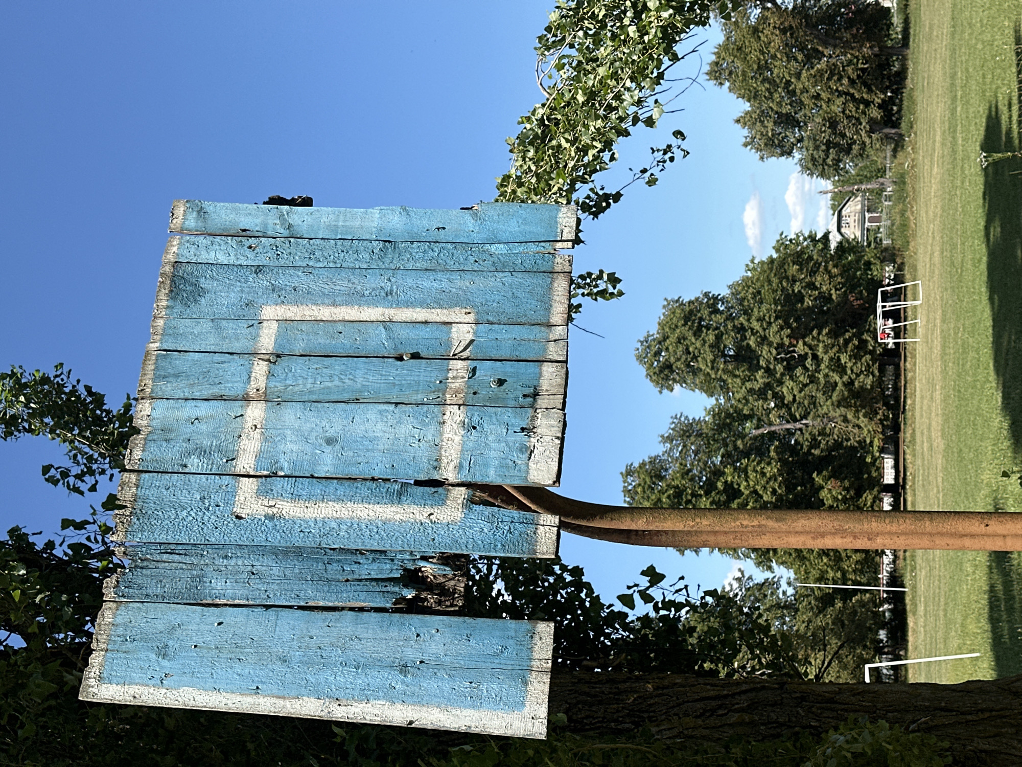 The image shows a dilapidated blue wooden basketball backboard with a white square painted on it, mounted on a rusted metal pole. The backboard is weathered and cracked, with some sections missing. It is positioned in an outdoor setting, with trees and a green field in the background. The sky is clear and blue. The field appears to be a sports field, with white goalposts visible in the distance. There is a house in the background, suggesting a rural or semi-rural location. The presence of the basketball backboard and goalposts implies a recreational area. The overall impression is one of neglect and disuse. It is not possible to determine the specific location in the city of Makiv, Ukraine based solely on this image.
