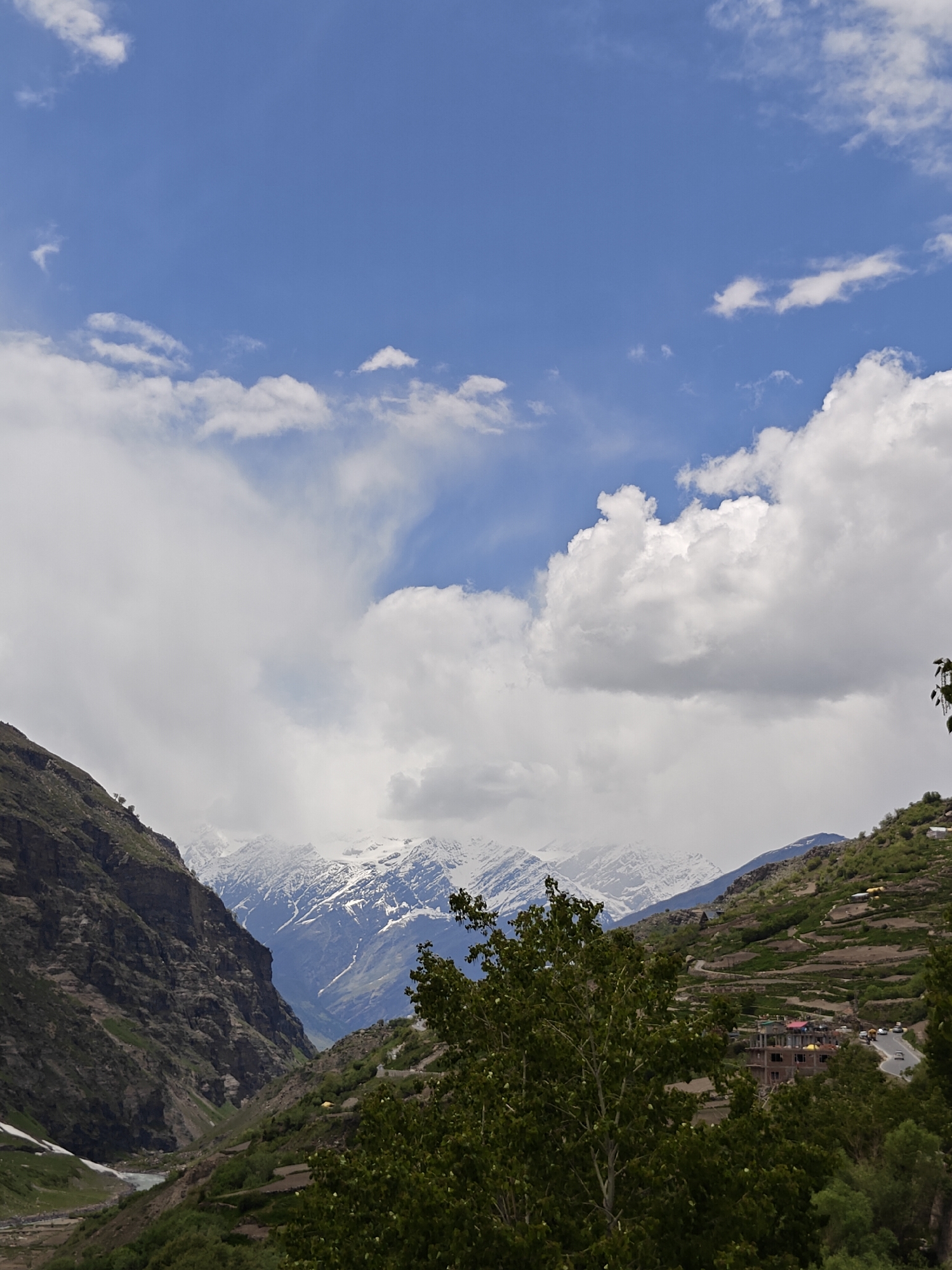 The image captures a mountainous landscape under a partly cloudy sky, likely in the vicinity of Yamling, India. Snow-capped mountains are visible in the distance, partially obscured by clouds. The foreground features green, forested slopes on either side of a valley. A river flows in the valley. On the right slope, terraced agriculture is apparent. A building is visible. The upper portion of the image shows a blue sky with scattered white clouds. The presence of terraced land suggests that this is a populated area.