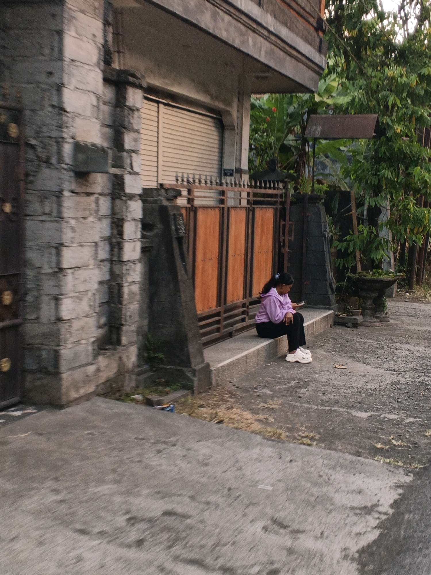 In Manggis, Indonesia, a young woman with dark hair is seated on a concrete step outside a building. She is wearing a purple hooded jacket and black pants, with white sneakers on her feet. The woman is looking down at her phone, which she is holding in her right hand. The building behind her has a stone facade and a large, retractable metal door. In front of the building is a decorative metal gate with orange wooden panels. To the right of the gate, there are trees and foliage, and a concrete planter. The ground in front of the building is paved with asphalt. The scene appears to be outdoors, likely during the daytime, with natural light illuminating the area.