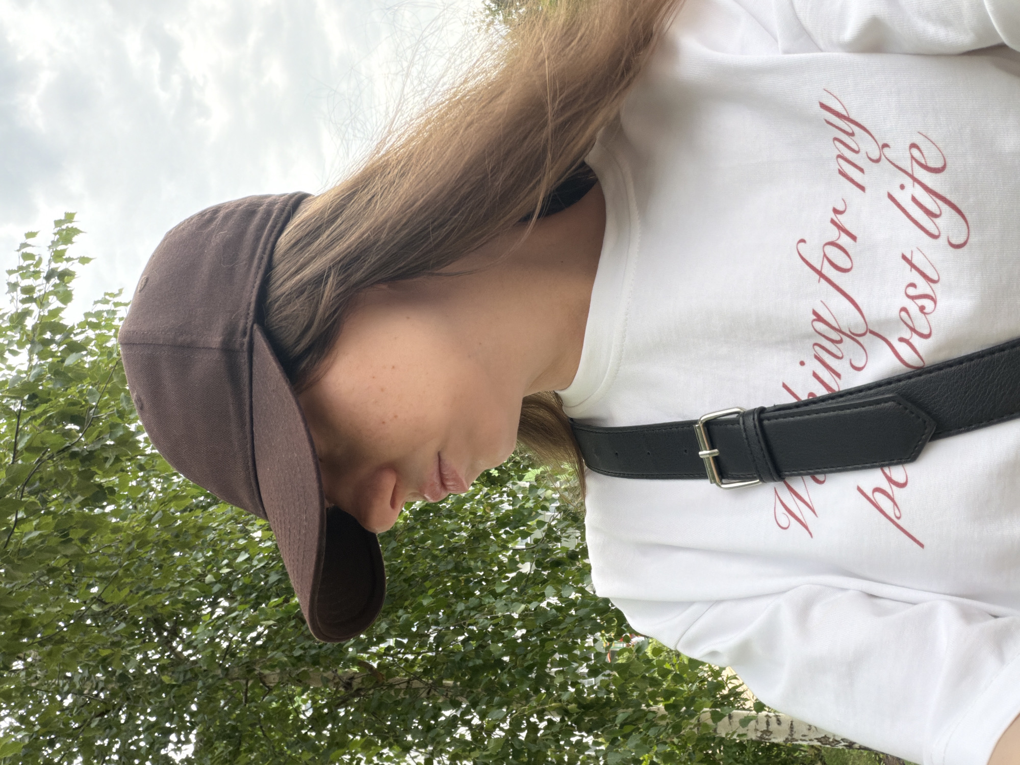 A woman wearing a brown baseball cap and a white t-shirt with red lettering stands outdoors. The t-shirt has text that reads "Working for my best life." She is also wearing a black crossbody bag. Behind her, there are green trees and a cloudy sky. The scene appears to be in a park or natural area within the city of Kyiv, Ukraine, possibly during daylight hours.