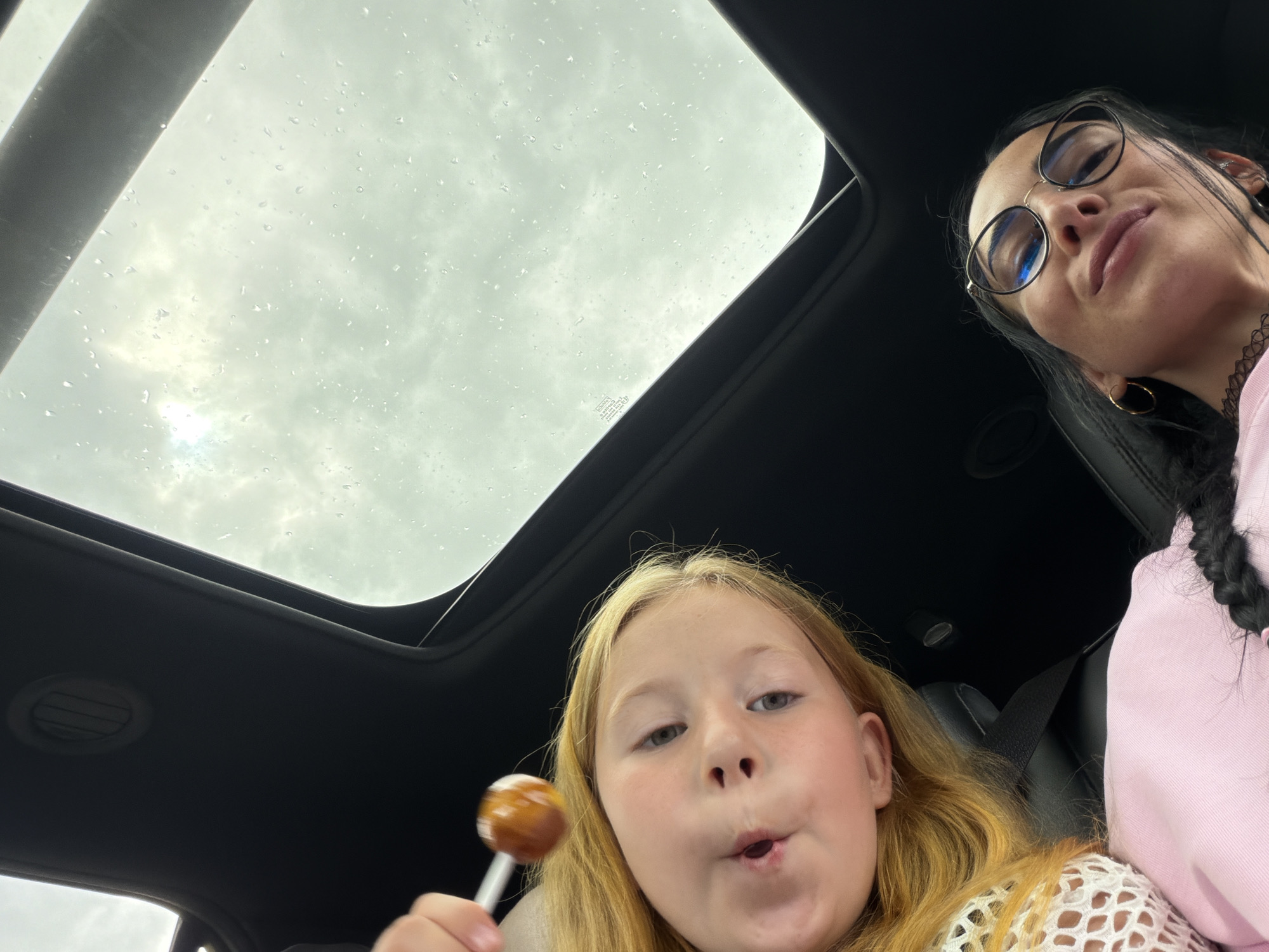 In Шолохово, Russia, two individuals are captured in a selfie from a low-angle perspective, seemingly inside a vehicle with an open sunroof. The sky above is overcast with visible raindrops on the glass. In the foreground, a young girl with blonde hair is holding a lollipop near her mouth. To her right, a woman with dark hair, wearing glasses and hoop earrings, is also looking towards the camera with a slight smile. The interior of the vehicle, likely a car, is visible with dark upholstery and a seatbelt.