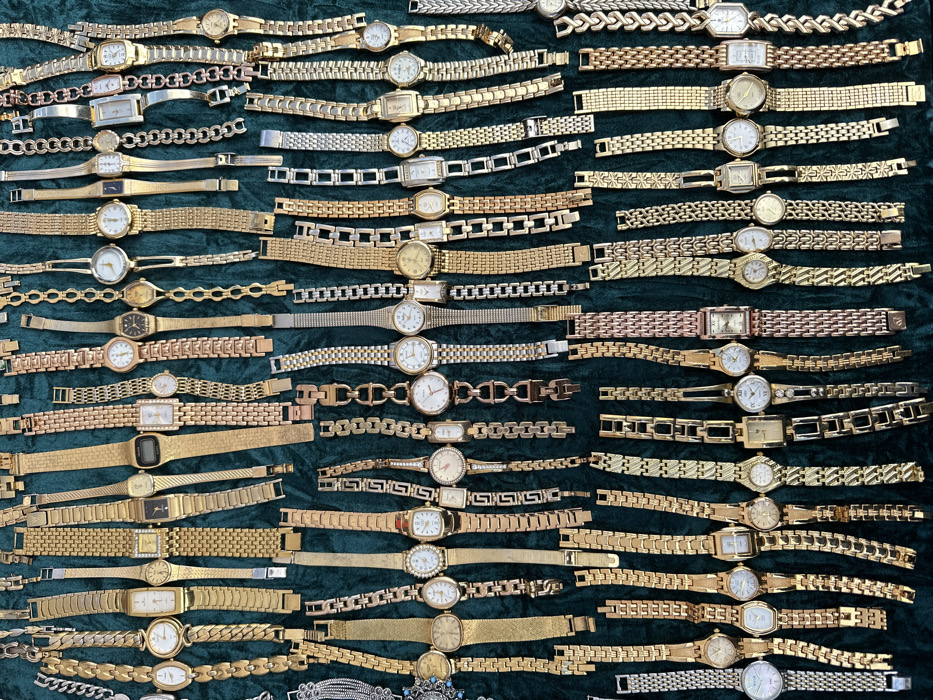 The image depicts approximately 150 to 180 diverse wristwatches, predominantly featuring gold-toned metal bands and cases, laid out on a dark green, possibly velvet or felt, fabric surface. The watches exhibit various designs, including round and rectangular faces with white, black, or gold dials, and a range of link bracelet styles. Some pieces incorporate silver-toned or rose gold elements. The collection is arranged in dense, overlapping rows, filling the entire frame. No individuals are present, and no actions or interactions are discernible. The scene is situated in the city of London, United Kingdom.