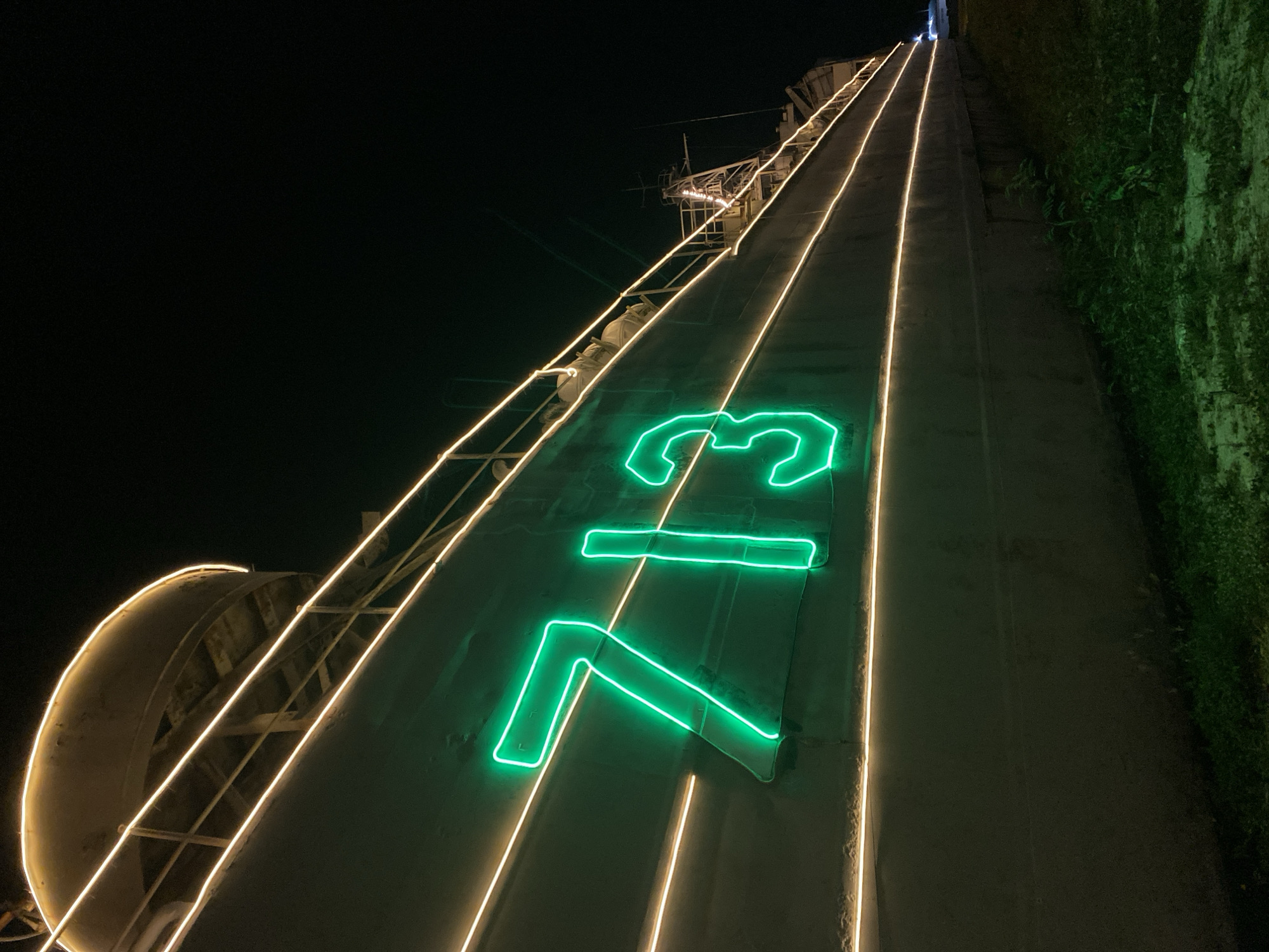 The image depicts a nighttime view of a structure in Baan Don Sai, Thailand, adorned with illuminated signage. The structure appears to be a tall, concrete incline, possibly a dam or retaining wall, flanked by vegetation on the right side. Two parallel lines of warm-toned lights run along the length of the incline, converging in the distance. A large, green neon sign displaying the number "73" is prominently featured on the structure's surface. The sky is dark, suggesting a night setting.