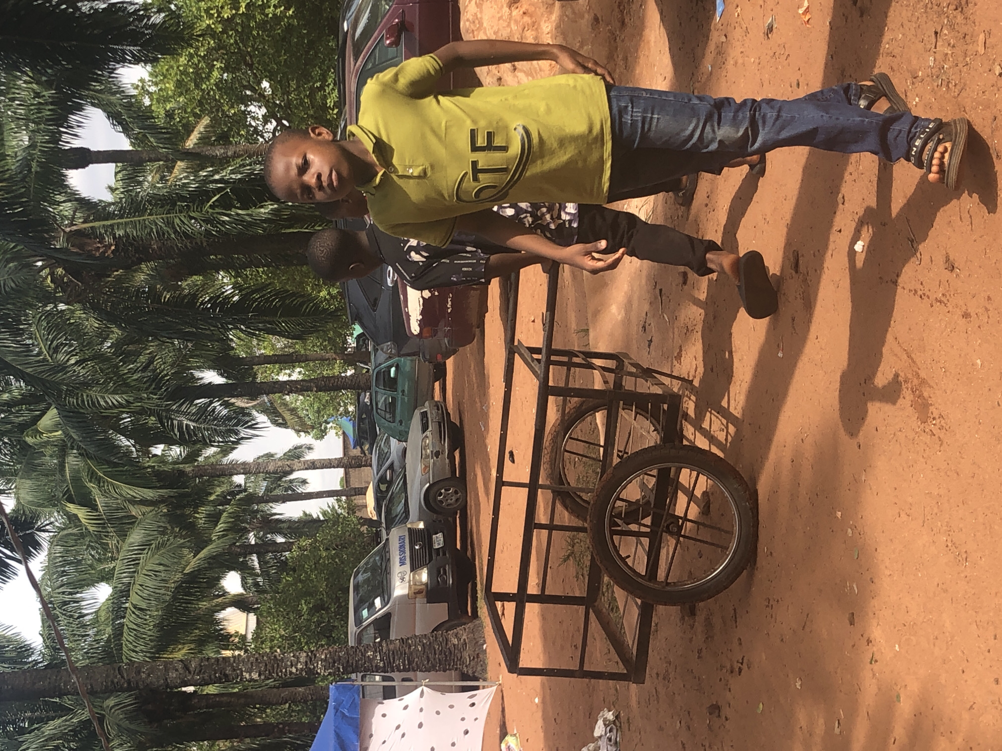 Two young boys stand on a red dirt ground in Nteje, Nigeria. One wears a yellow shirt and jeans, the other a black and white patterned shirt with black pants. They stand next to a metal-framed cart with bicycle wheels. Behind them, a row of cars and vans are parked, and tall palm trees line the scene's left side.