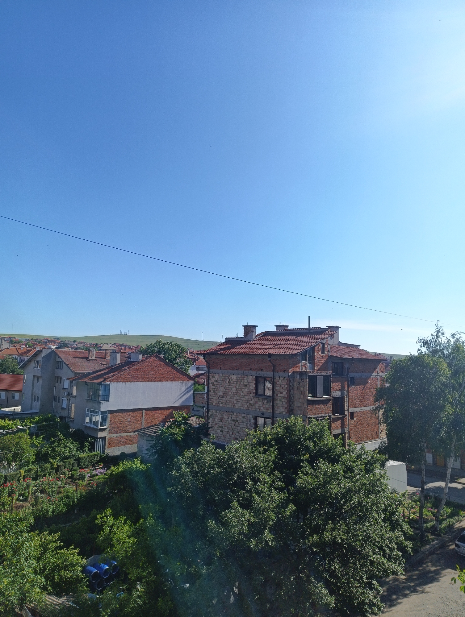 The image depicts a view from an elevated position, looking out over a residential area with houses under construction or renovation, characteristic of a developing neighborhood in Karnobat, Bulgaria. The sky is clear blue, with the sun high overhead, suggesting a daytime setting. Several buildings are visible, some with brick facades and unfinished roofs, indicating ongoing construction work. Red-tiled roofs are prevalent on completed structures. Green trees and gardens are interspersed throughout the area, adding a touch of nature to the urban landscape. In the distance, rolling hills are visible, with some communication towers or industrial structures dotting the horizon. The overall impression is of a sunny day in a Bulgarian town, with a mix of older and newer constructions.