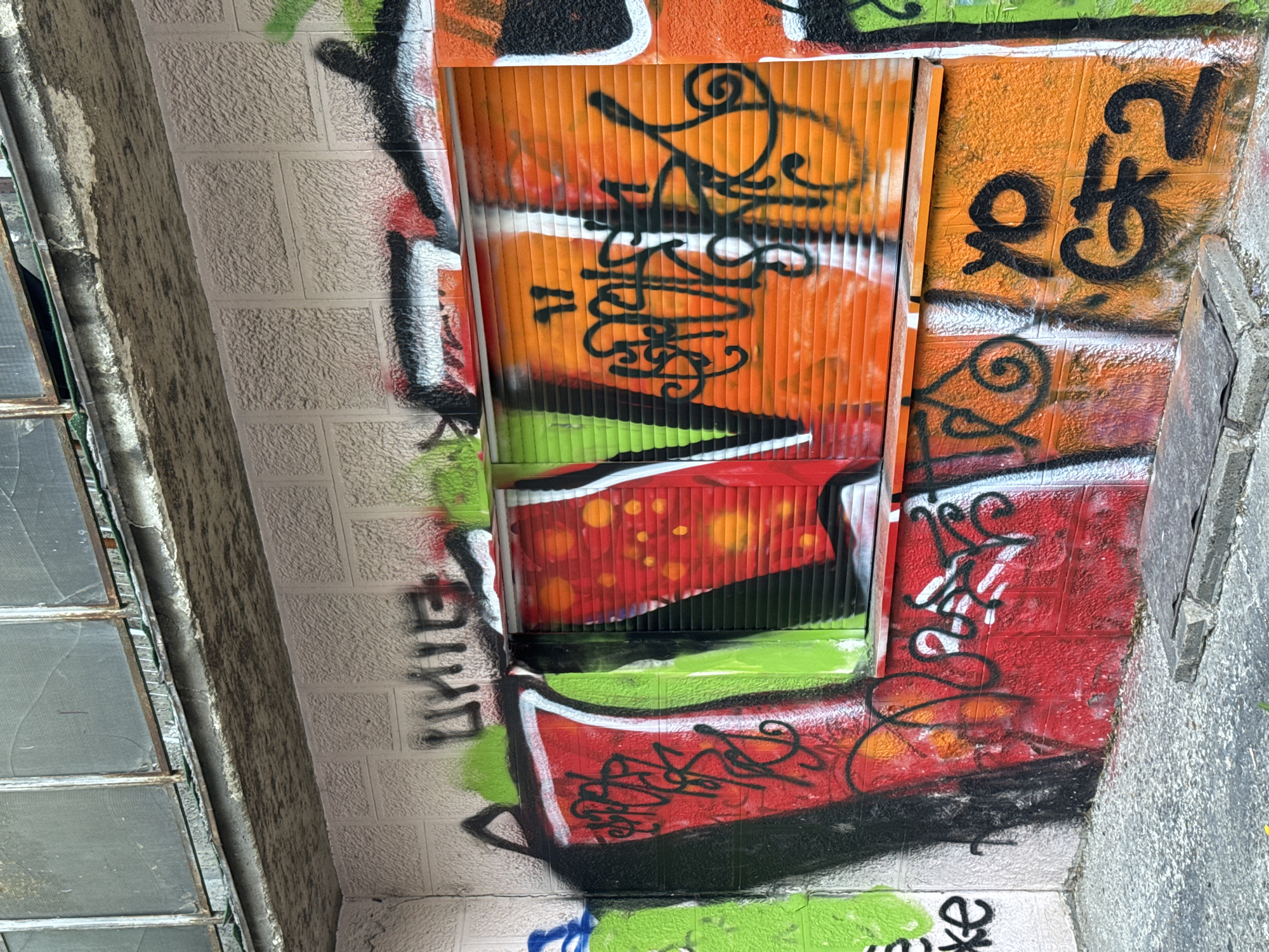 The image depicts an exterior wall covered in graffiti, located in Belgrade, Serbia. The wall is constructed of light-colored brick or concrete blocks, heavily painted with various colors, predominantly orange, red, green, and black. Much of the paint appears to be applied in a layered fashion, indicating multiple instances of graffiti art or tagging.
A prominent feature is a corrugated metal door or panel, also covered with graffiti. This panel is painted orange and green, with black lettering and designs obscuring most of the original color.
To the left, a portion of a window frame is visible. The windowpanes appear to be cracked and dirty.
To the right, there is a concrete or stone edge.