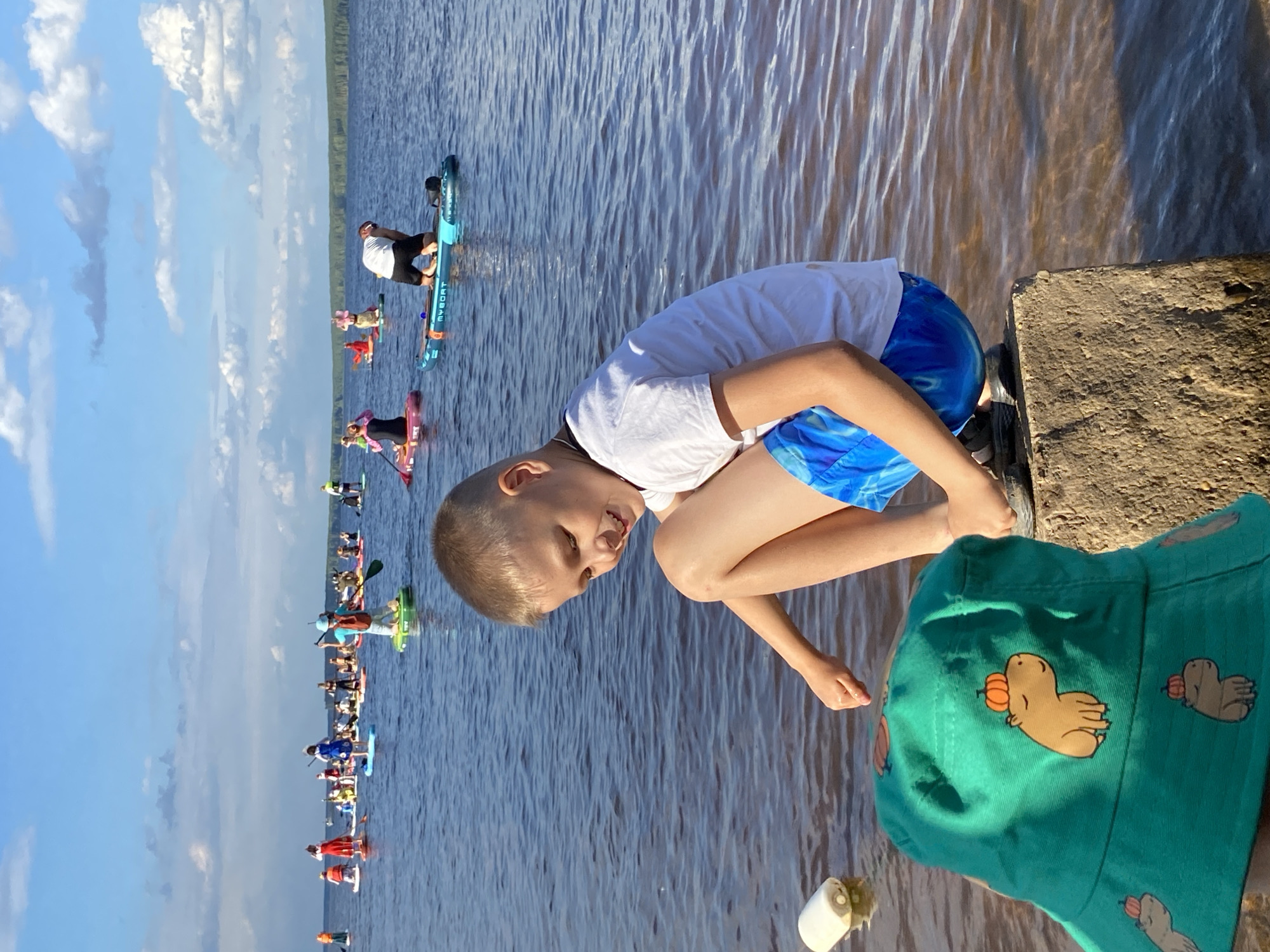 A group of people are paddleboarding on a large body of water under a bright, cloudy sky. The scene is likely taking place in the late afternoon, given the warm lighting and shadows. The water is calm, with ripples reflecting the sunlight. In the foreground, a young boy wearing a white t-shirt and blue patterned shorts is crouched down, smiling and looking towards the camera. He is on a concrete embankment to the right of the frame. Next to him, the top of another person's head and shoulders are visible, wearing a green bucket hat adorned with cartoon capybara and pumpkin prints.
In the background, a procession of paddleboarders stretches across the water. Many are standing on their boards, some with paddles in hand. The boards are of various colors, including blue, pink, and green. The paddleboarders appear to be participating in an organized event or a group outing. The distant shore is lined with trees.
The overall mood of the image is cheerful and active, with the focus on the boy's evident enjoyment and the communal activity on the water. The clear sky and calm water suggest pleasant weather conditions suitable for water sports.