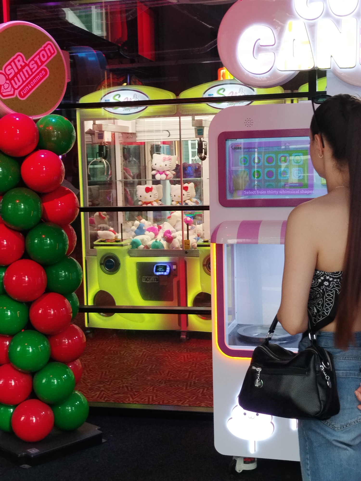 A woman stands in front of a claw machine game in an arcade. She is wearing a black bandana and blue jeans, and is carrying a black purse. The claw machine is brightly lit, with plush toys visible inside. In the background, a sign reads "SIR WINSTON FUN & GAMES". The arcade appears to be indoors, with a patterned carpet on the floor.