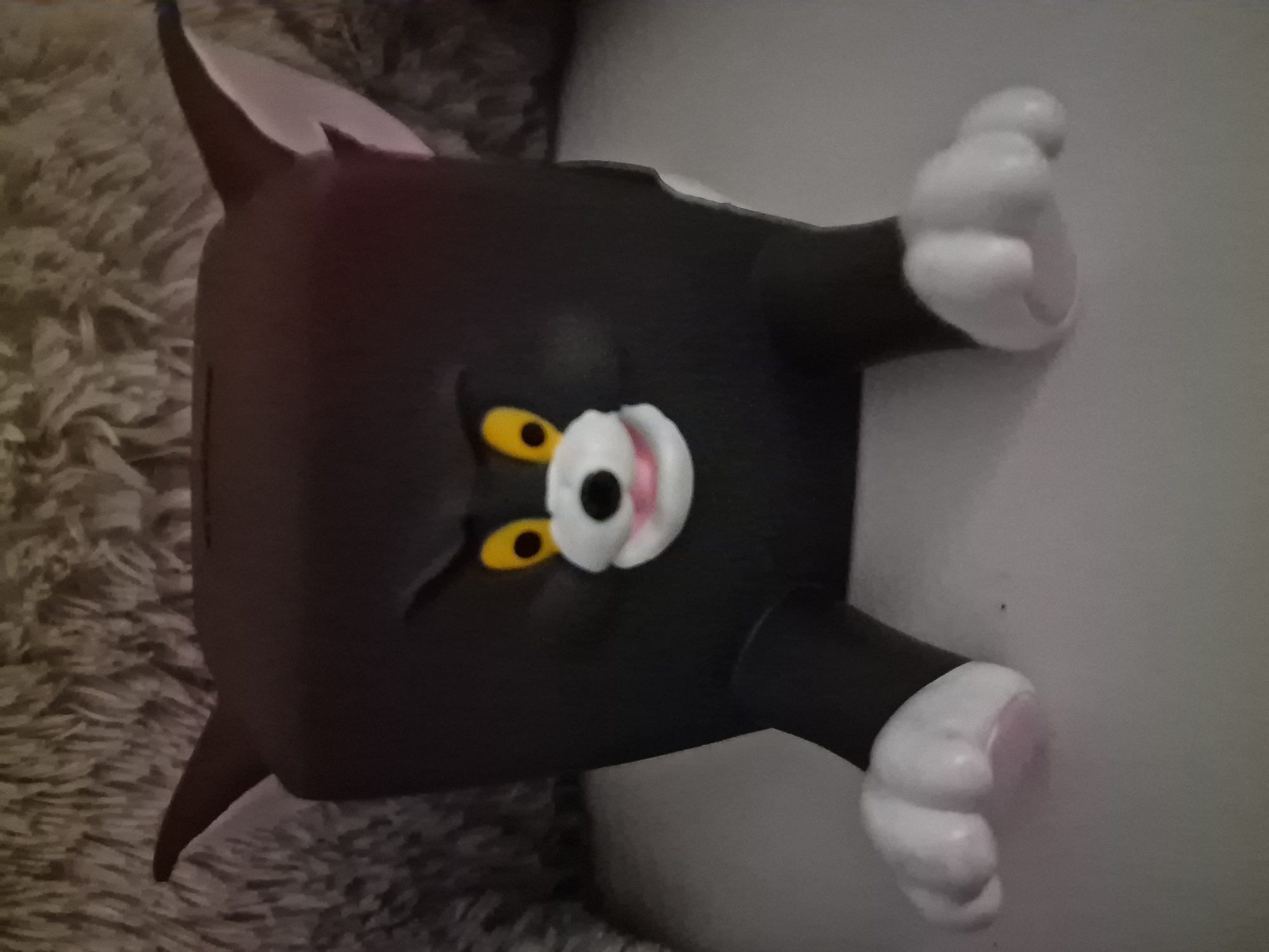A dark gray, square-shaped piggy bank designed to resemble the cartoon character Tom the cat is the central subject. The piggy bank has yellow eyes with black pupils, a white muzzle with a black nose and pink mouth, and white paws with pink pads. It is positioned on a light gray surface, possibly a table or shelf. Behind the piggy bank, a textured, shaggy, light gray surface is visible, likely a blanket or rug. The overall lighting suggests an indoor setting. The image does not provide any information that can identify its location in the city of Kursavka, Russia.
