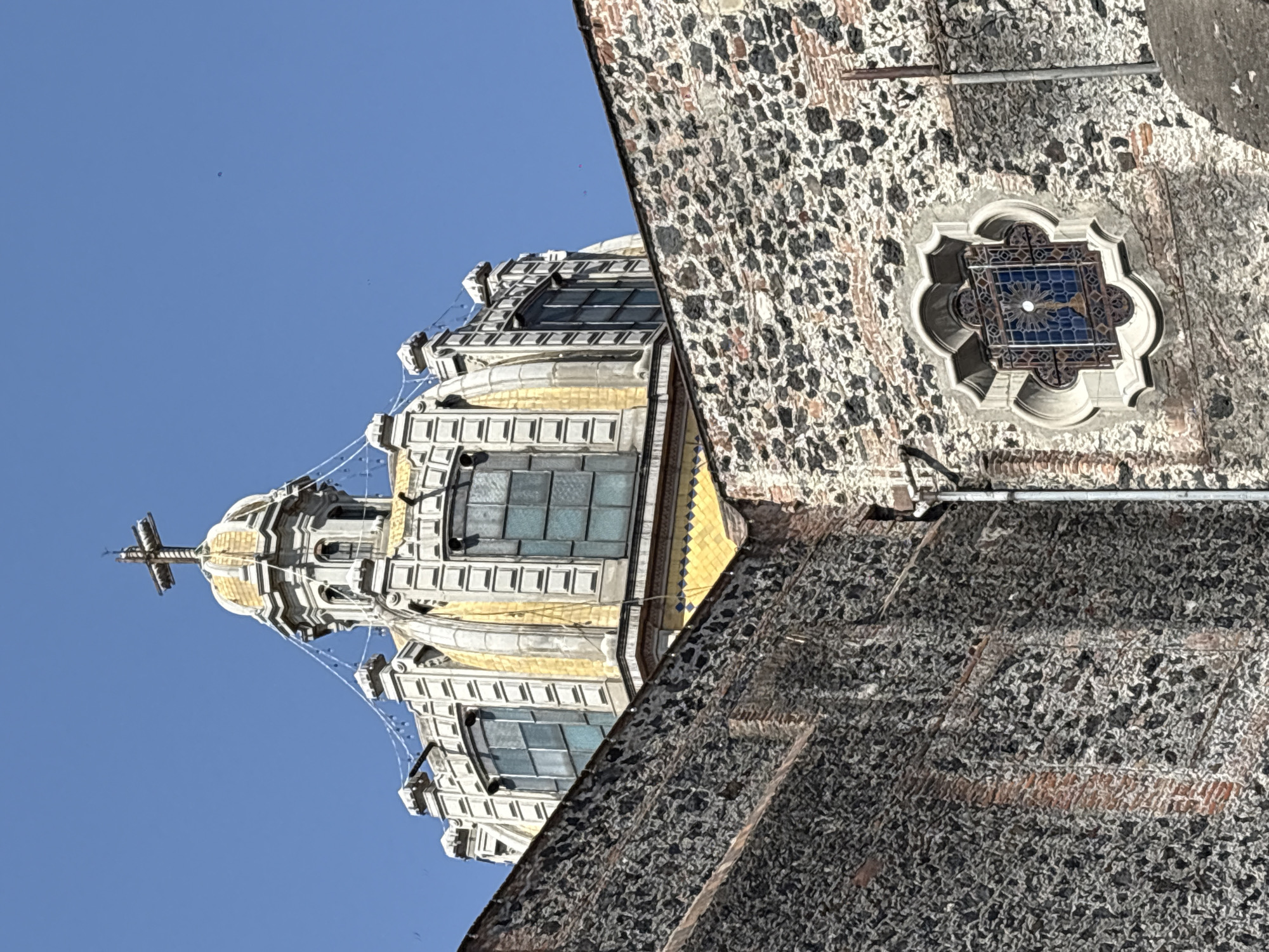 The image shows a low-angle view of a building in Mexico City. The building appears to be a church or cathedral with a prominent dome topped by a cross. The dome is multi-tiered, with windows and decorative elements. The walls are made of rough, textured stone. A small, decorative window with a blue interior is visible on the side of the building. The sky is clear and blue.