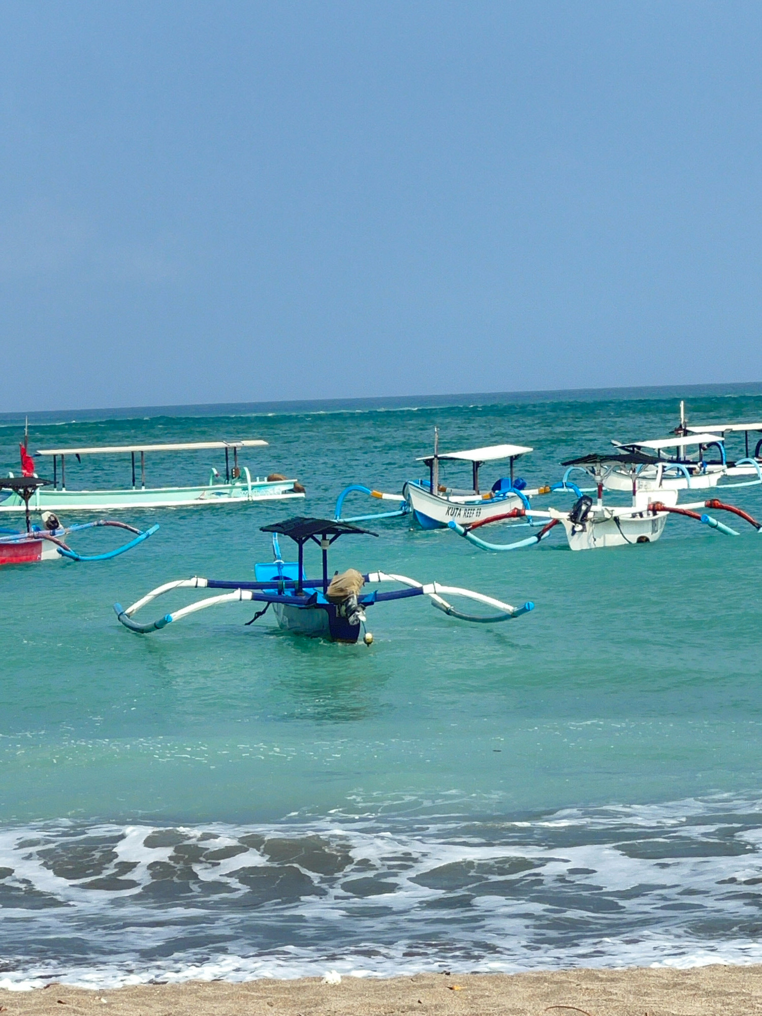 The image shows a beach in Kuta, Indonesia, with several Balinese outrigger boats (jukungs) floating on the turquoise water. The sky is a clear, pale blue. The boats are of various sizes and colors, with some featuring canopies for shade. One boat in the foreground is blue and white with a dark canopy and an outboard motor. Another boat in the middle ground is white with blue trim and a red stripe along its side. In the background, more boats are visible, and the ocean extends to the horizon. The shoreline is a sandy beach with some small waves breaking.