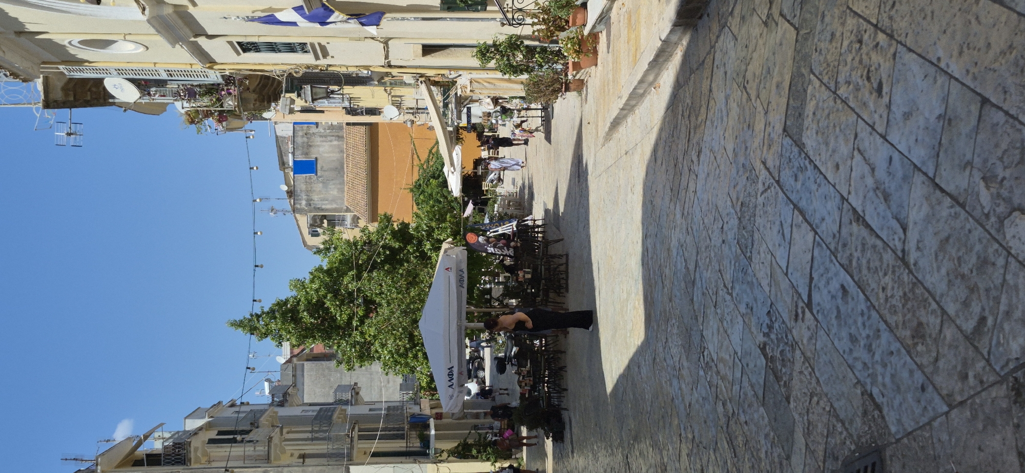 The image depicts a street scene in Corfu, Greece, likely within the historic Old Town. The perspective is from a slightly elevated viewpoint looking down a cobblestone street lined with buildings. In the foreground, the textured cobblestones cast long shadows from the overhead sun.

Several people are visible. In the middle ground, a woman in a black dress stands near a white umbrella, possibly an outdoor cafe. Further down the street, a couple in white robes walks away from the camera. Other individuals are present, some seated at tables under the umbrella and others walking in the distance.

The environment is characteristic of Mediterranean architecture, with multi-story buildings in shades of cream, yellow, and a muted orange. Balconies with wrought-iron railings and shuttered windows are visible. A large green tree stands prominently in the mid-ground, providing shade. String lights are strung across the street overhead. In the background, various buildings, antennas, and satellite dishes indicate a populated urban setting. The sky is a clear, bright blue, suggesting a sunny day. The presence of outdoor seating and umbrellas suggests a commercial area, likely a pedestrian zone with shops or cafes.