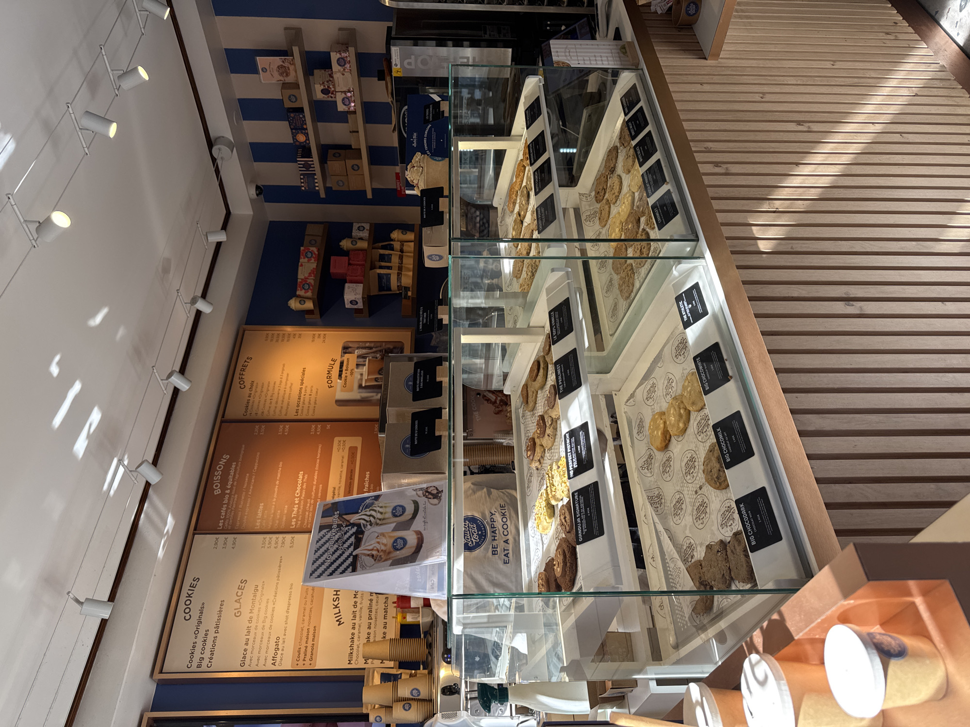 The image depicts the interior of a bakery, identified as "Laura Todd Cookies," located at 38 Rue de la Monnaie in Lille, France. The scene focuses on a display counter filled with various cookies and pastries. The counter is illuminated by track lighting, and sunlight streams through a window, casting shadows on the wooden paneling of the counter.
In the foreground, a glass display case showcases an assortment of cookies on white paper liners. Labels beneath the cookies identify specific flavors such as "GIANDUJA SIGNATURE," "THE PERFECT PISTACHE," "THE PRALINET," "BIG CHOCOLMILK," "BIG CHOCOLDARK," "BIG CHOCOWHITE," "CARAMELT," "CHOCOLAT PARISIEN," and "WHITE DELICE." Some of the cookies are round and thick, while others are flatter and more oblong.
Behind the display case, a menu board with sections for "COOKIES," "GLACES," "MILKSHAKE," "BOISSONS," "Les cafés bio & équitables," "Les cafés," "Les lattes," "Les Thés et Chocolats," and "FORMULE" is visible. A folded promotional sign featuring images of ice cream cones is placed on the counter.
To the left, stacked paper cups and lids are arranged on a wooden shelf. Further back, shelves mounted on a wall with blue and white stripes hold boxes of cookies and other packaged goods. A coffee machine and a small refrigerator are also partially visible.
No people are clearly visible in the scene, suggesting the establishment may be temporarily unstaffed or the individuals are out of frame. The overall environment is clean and well-lit, typical of a retail food establishment.