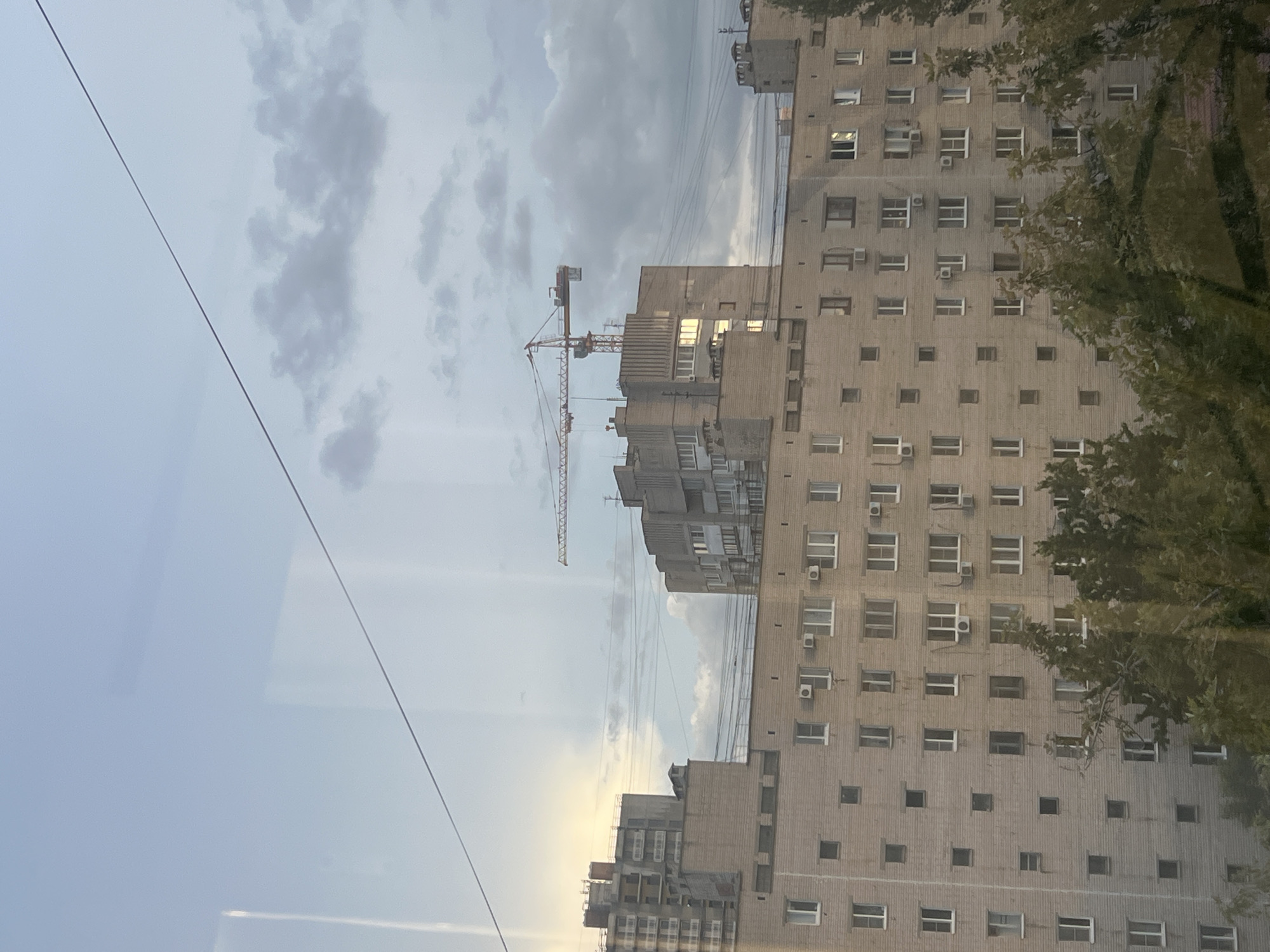 The image shows a cloudy sky above several large apartment buildings, with a construction crane visible in the distance. The buildings are made of light-colored brick and have many windows. Trees are visible in the foreground, partially obscuring the lower floors of the buildings. The scene is likely a residential area in Volgograd, Russia, given the architectural style of the buildings and the presence of a construction crane, which is common in urban development. There are no people visible in the image. The overall atmosphere is somewhat overcast, with the light suggesting either early morning or late afternoon. The image appears to have been taken from a window, as evidenced by reflections on the glass.