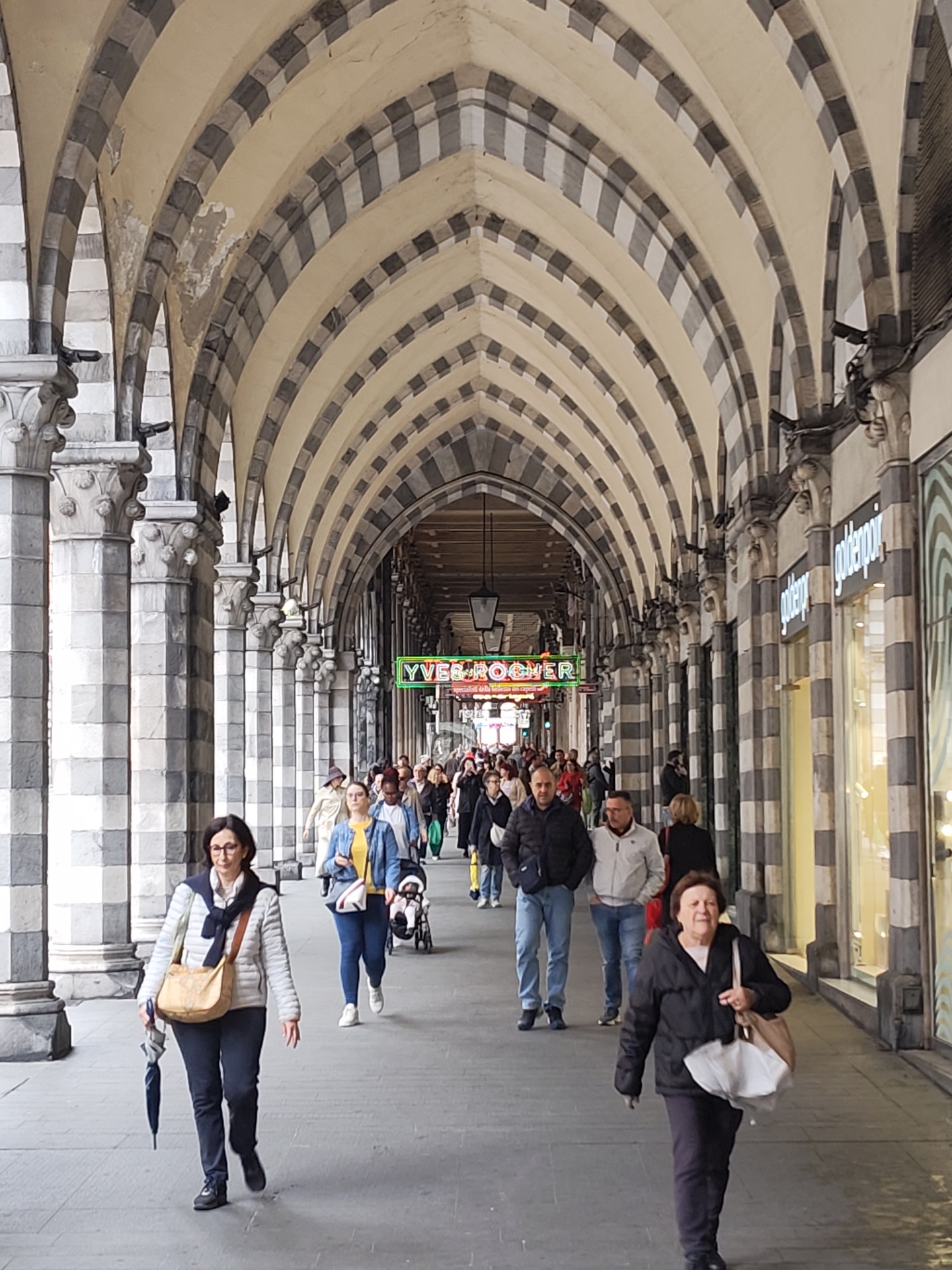 The image depicts a covered walkway, likely in the city of Genoa, Italy. The architecture features repeating archways with alternating dark and light stone or paint. A crowd of people is walking down the walkway. Among the people are two women in the foreground and men and women further down the walkway. A neon sign reading "Yves Rocher" is visible in the distance above the crowd. Retail shops line the right side of the walkway. The day appears to be overcast.