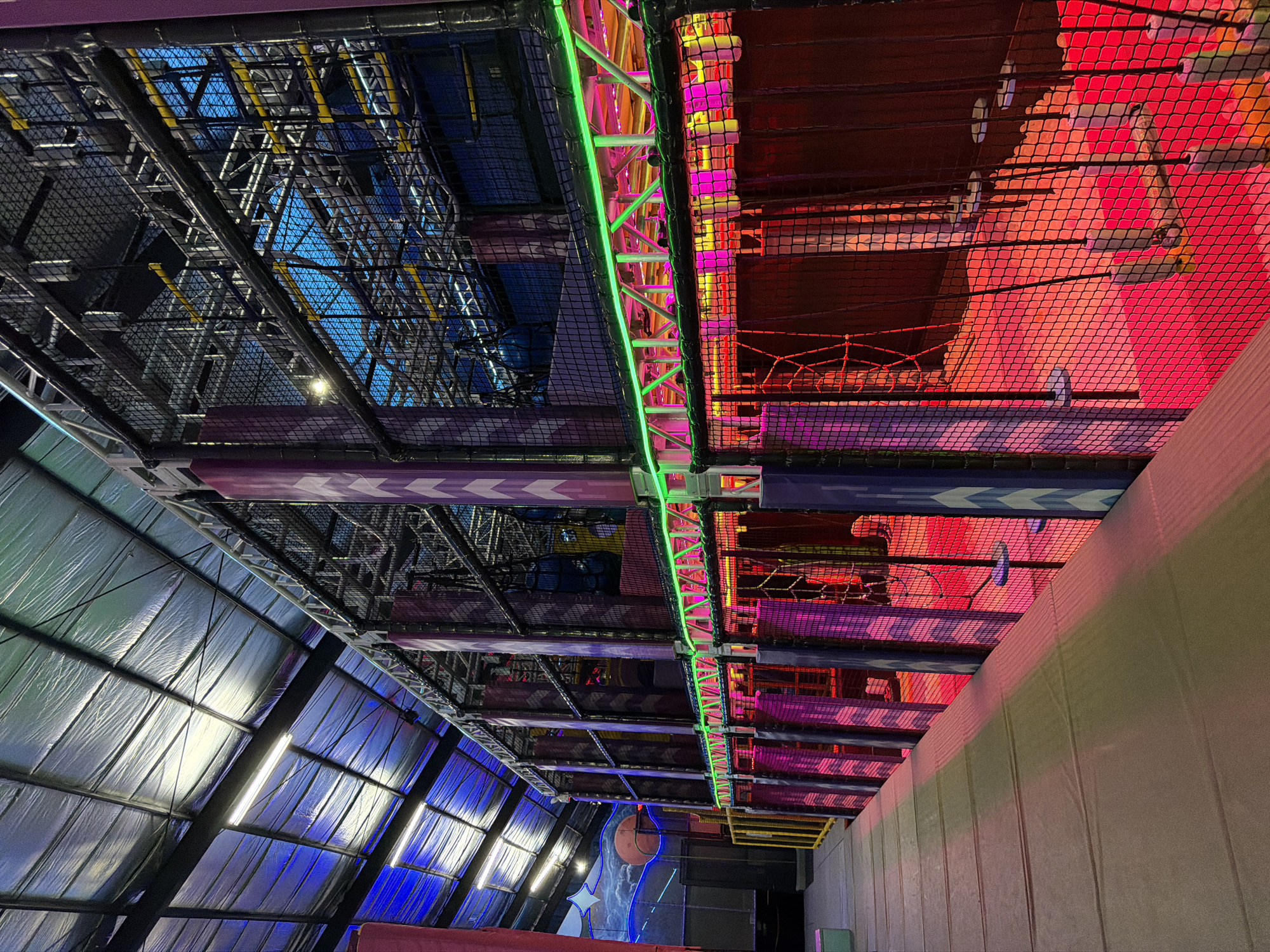 The image depicts the interior of what appears to be an indoor adventure park in Denpasar, Indonesia. The camera angle is pointed upwards, capturing the ceiling structure and elements of the park's design.
To the left of the frame, a large, sloped metallic wall is visible, illuminated with blue-toned lighting. Fluorescent lights are mounted on the wall. A basketball court area is visible below.
Overhead, a complex network of metal framework and netting creates a suspended play area. Sections of the structure are illuminated with different colored lights, including green, pink, and red. Some areas are a dense maze-like construction, enclosed by netting.
