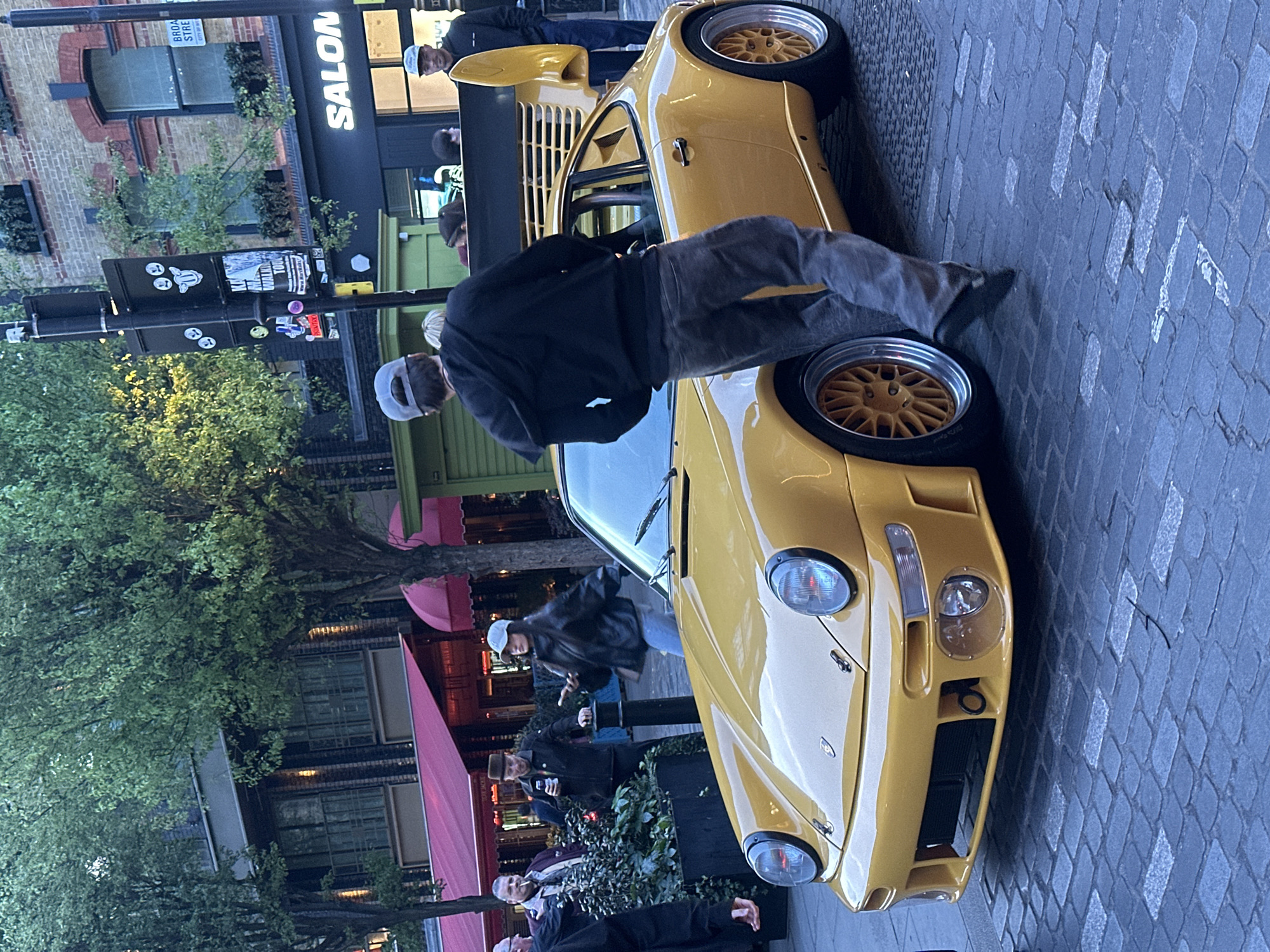 This evening street scene, set in London, United Kingdom, features a bright yellow sports car, resembling a classic Porsche 911 model, parked on a cobblestone street. Several individuals are gathered around the car, with one person in a black jacket and grey cap leaning over the front hood, seemingly admiring or inspecting the vehicle. Another person in a cap stands by the car's rear. The car itself has striking gold multi-spoke wheels and prominent round headlights.
The background reveals a bustling urban environment. Trees with green foliage line the street, and commercial buildings with large windows and awnings are visible. One prominent building displays the word "SALON" in white letters on a dark facade. A street sign above this building partially reads "BROAD STRE" (likely "Broad Street"). A lamppost near the car is adorned with various stickers, including one that partially reads "KATE WAITED FOR..." and another red sticker with "BORED". The subdued lighting suggests dusk or early evening, with interior lights from shops casting a glow.
